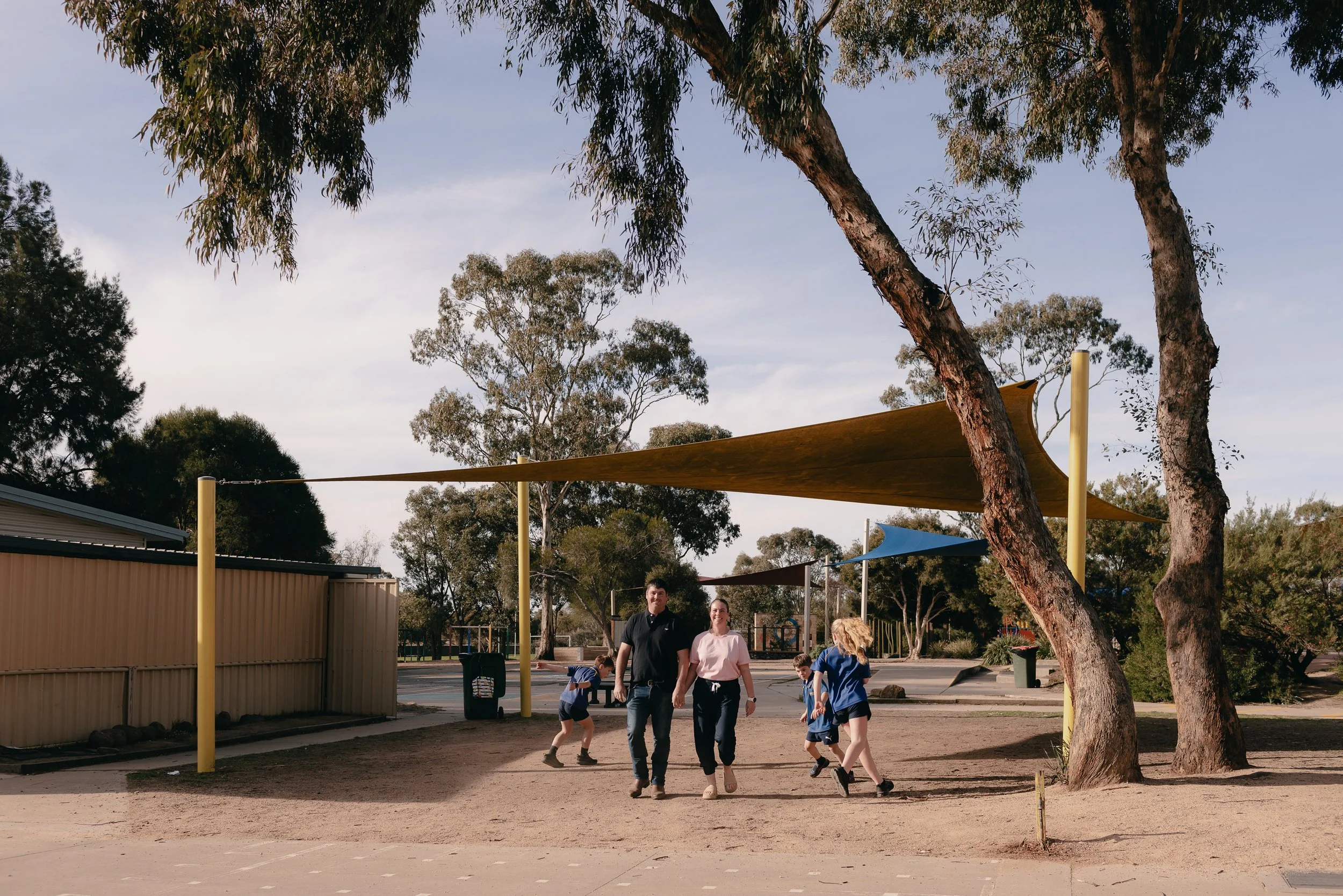 Adults and children walking in a playground area with shade sails and trees.