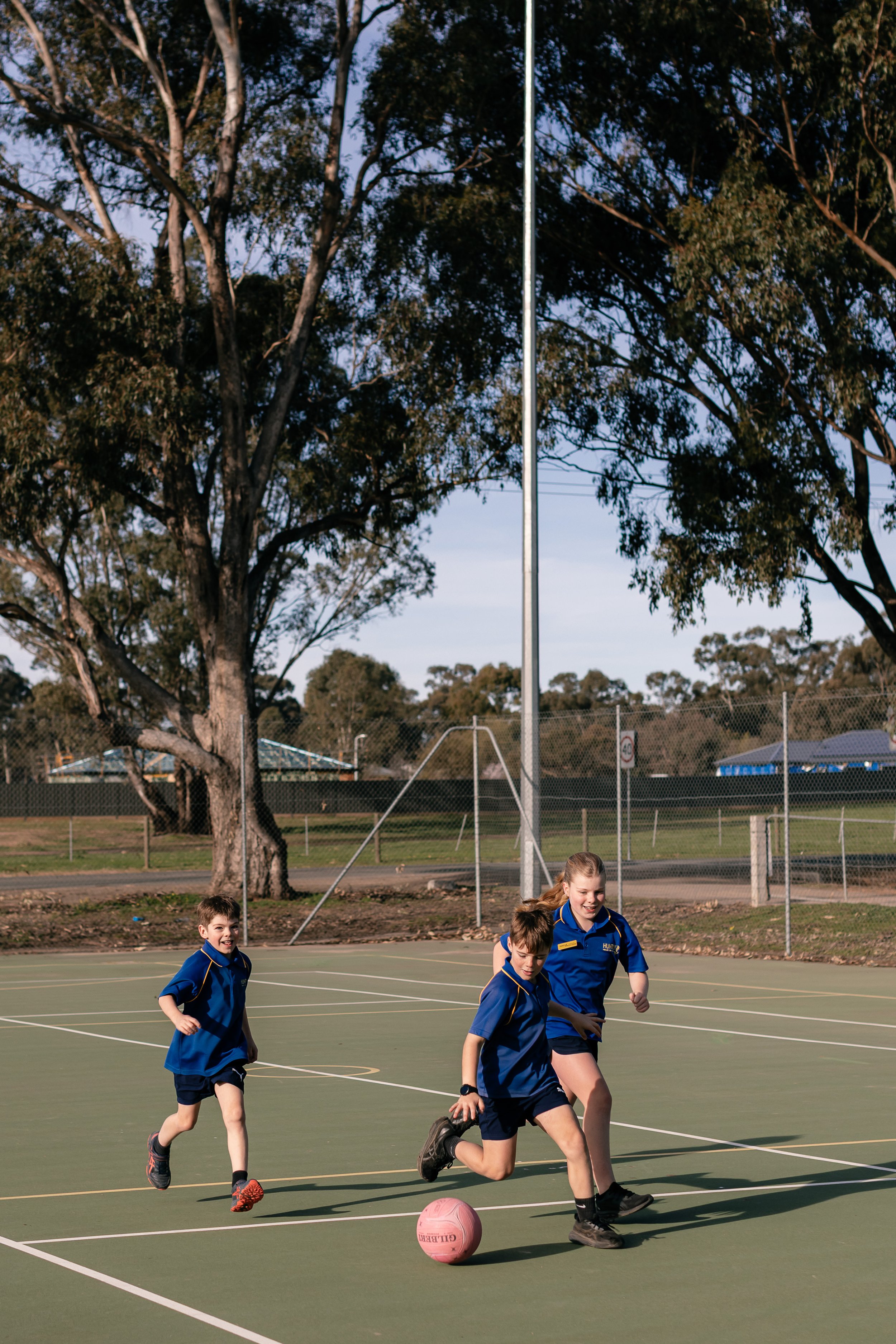 Three children playing soccer outdoors on a green court, with large trees and a chain-link fence in the background, during daylight.