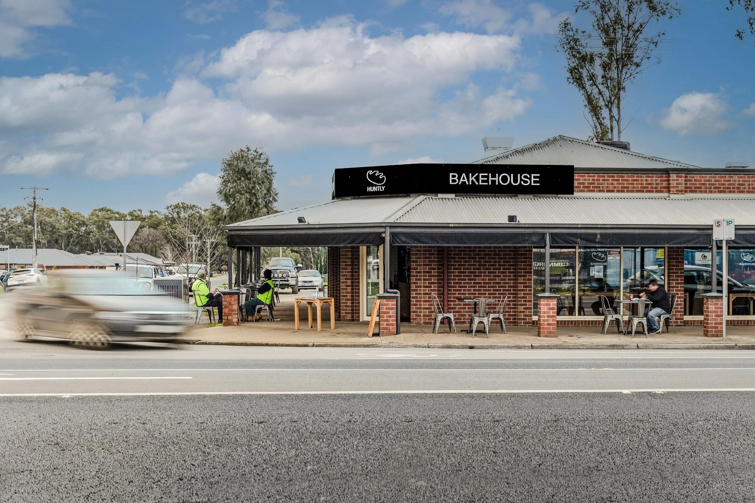 A brick building with a black sign that says 'Bakehouse' and the Hitchy logo, with outdoor seating and people sitting at tables across the sidewalk. There are cars passing by on the road, and trees in the background under a partly cloudy sky.