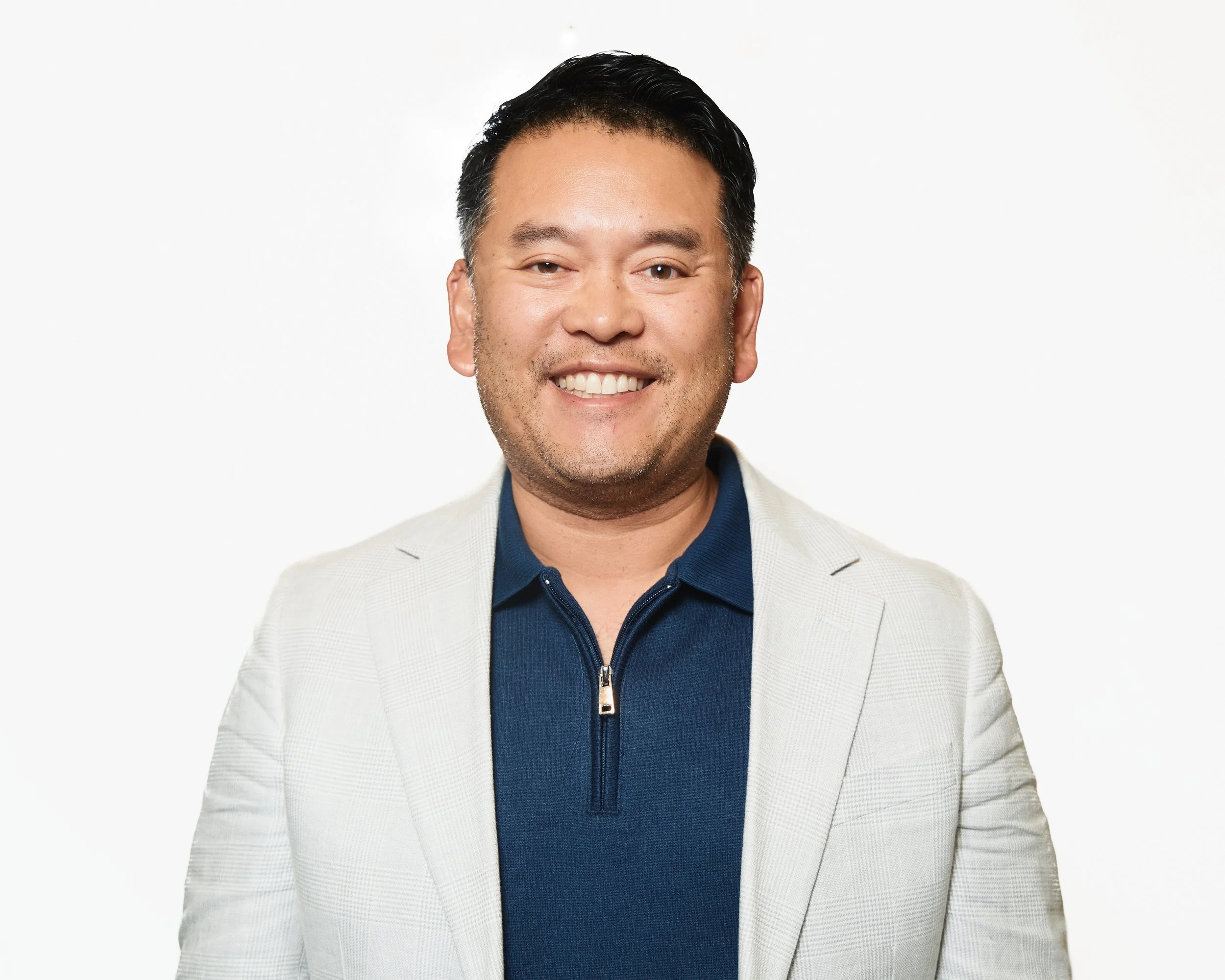 A smiling man with short dark hair, wearing a light-colored blazer and a dark blue zip-up shirt, standing against a plain white background.