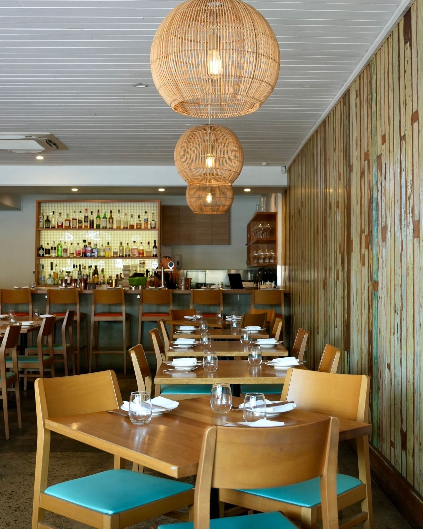Interior of a restaurant with wooden tables and chairs, glassware and plates set on the tables, a bar with bottles in the background, and hanging wicker pendant lights.