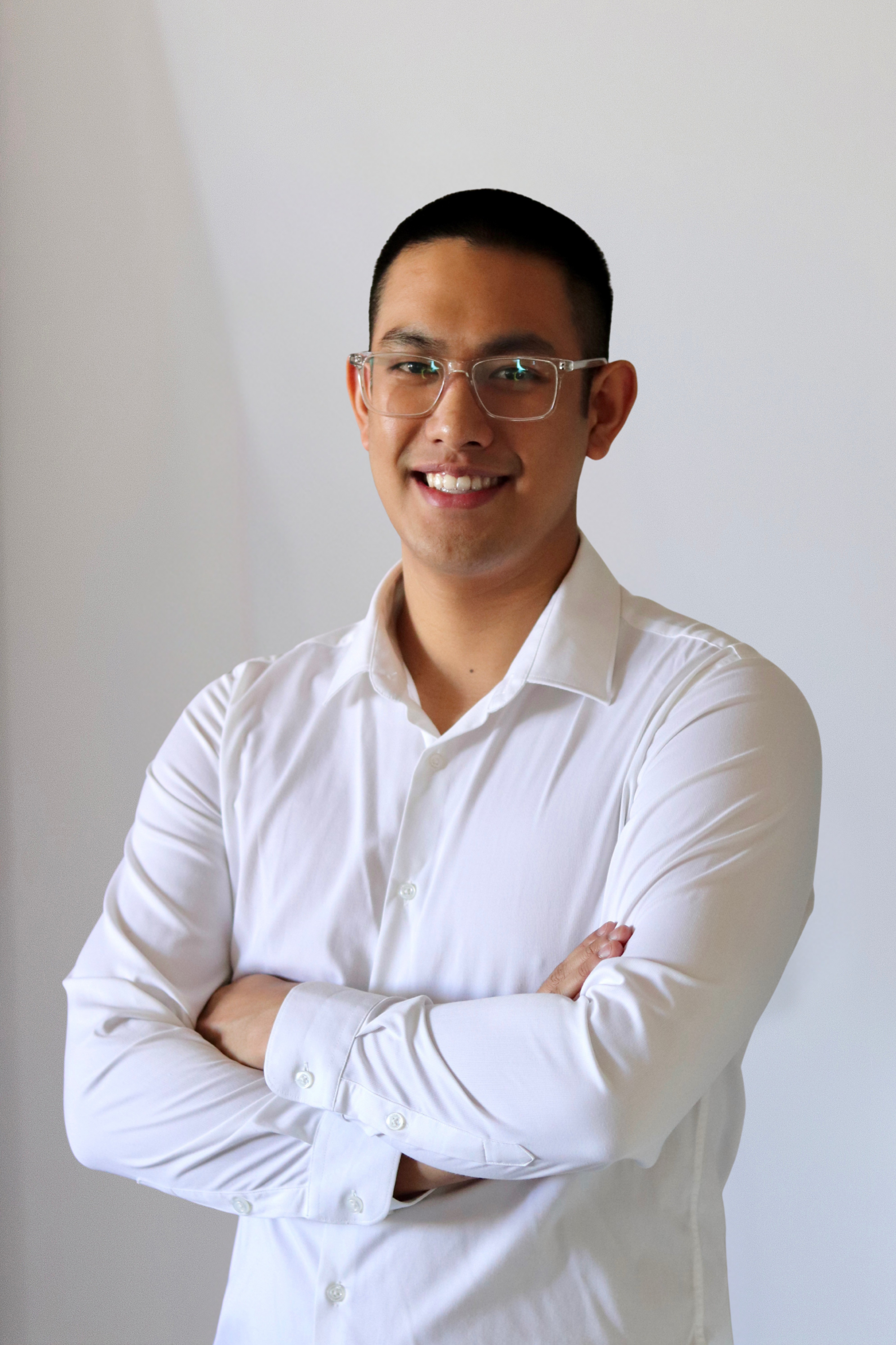 A young man wearing glasses and a white button-up shirt, standing with arms crossed and smiling against a plain light-colored background.