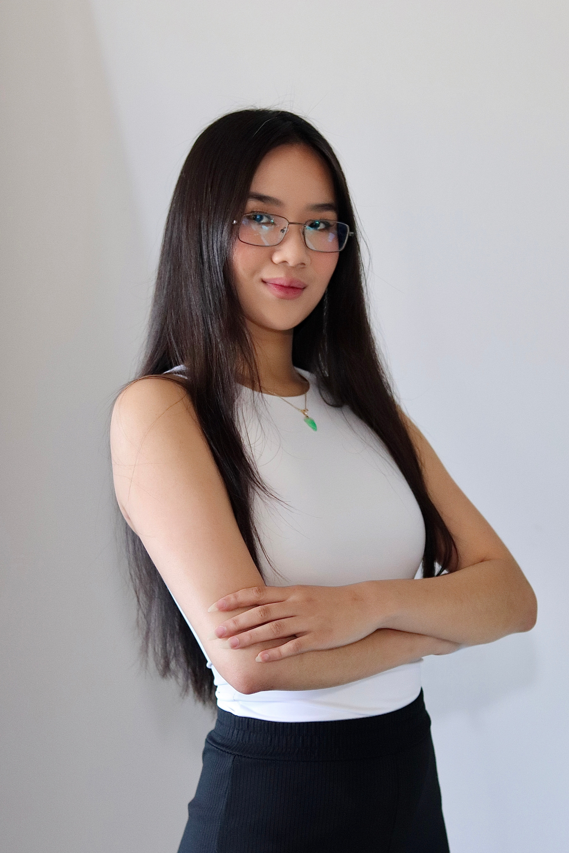 Young woman with long dark hair, glasses, and a white sleeveless top, crossing her arms and looking at the camera against a plain background.