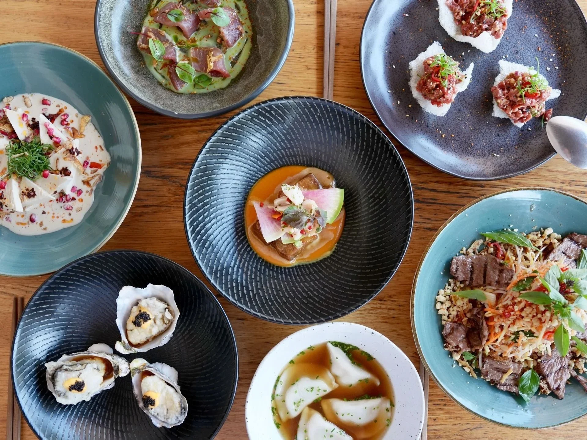 A variety of Asian dishes on a wooden table, including oysters, beef tartare, soup, rice with beef and vegetables, and assorted appetizers.