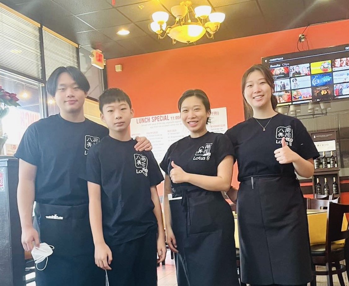Four restaurant staff members wearing black shirts with 'Lotus 8' logo, standing inside a restaurant with a red wall and a menu board in the background, smiling and posing for the photo.