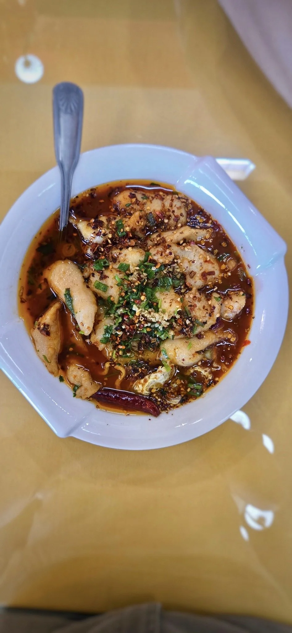 A bowl of Sichuan, shui zhu yu, or spicy poached fish filet,  in spicy chili oil topped with chopped green onions, mala spice, cilantro and red pepper flakes, with a spoon inside, on a glass table.