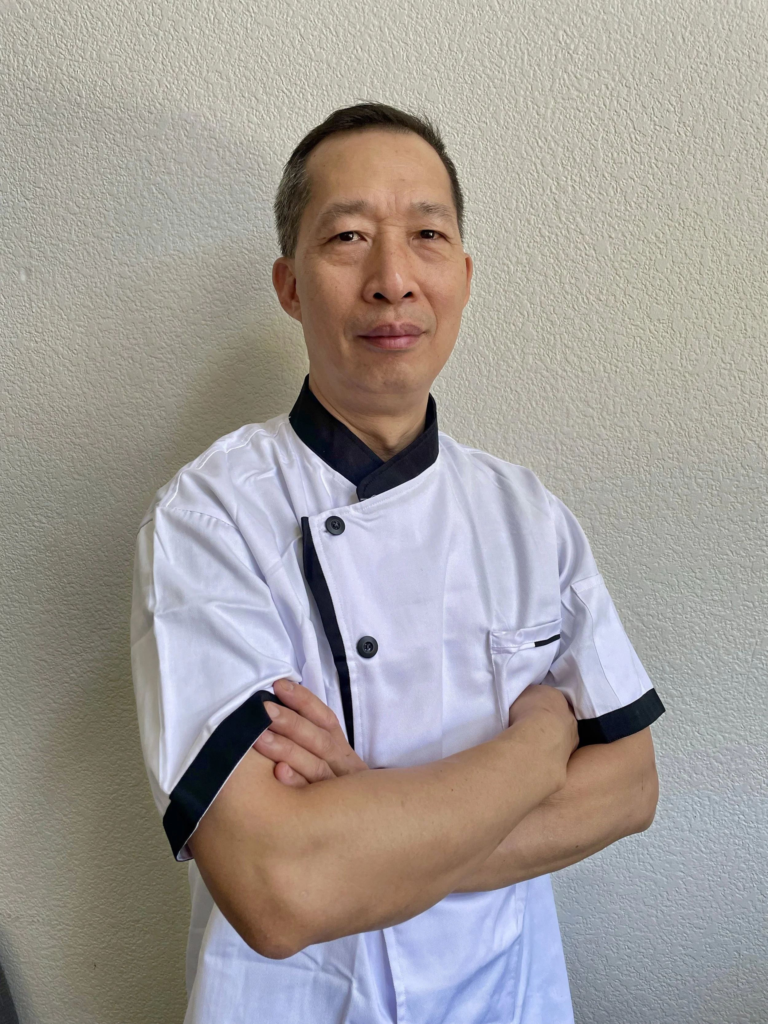 A middle-aged man wearing a white chef's coat with black accents, arms crossed, standing against a plain off-white wall.
