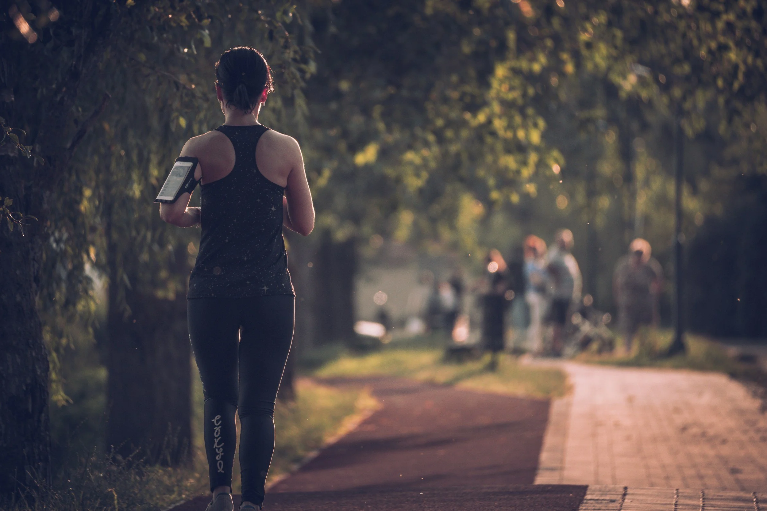 A woman running or walking on a park path, wearing athletic clothing and a phone arm band. Return to running. Bounce back after pregnancy