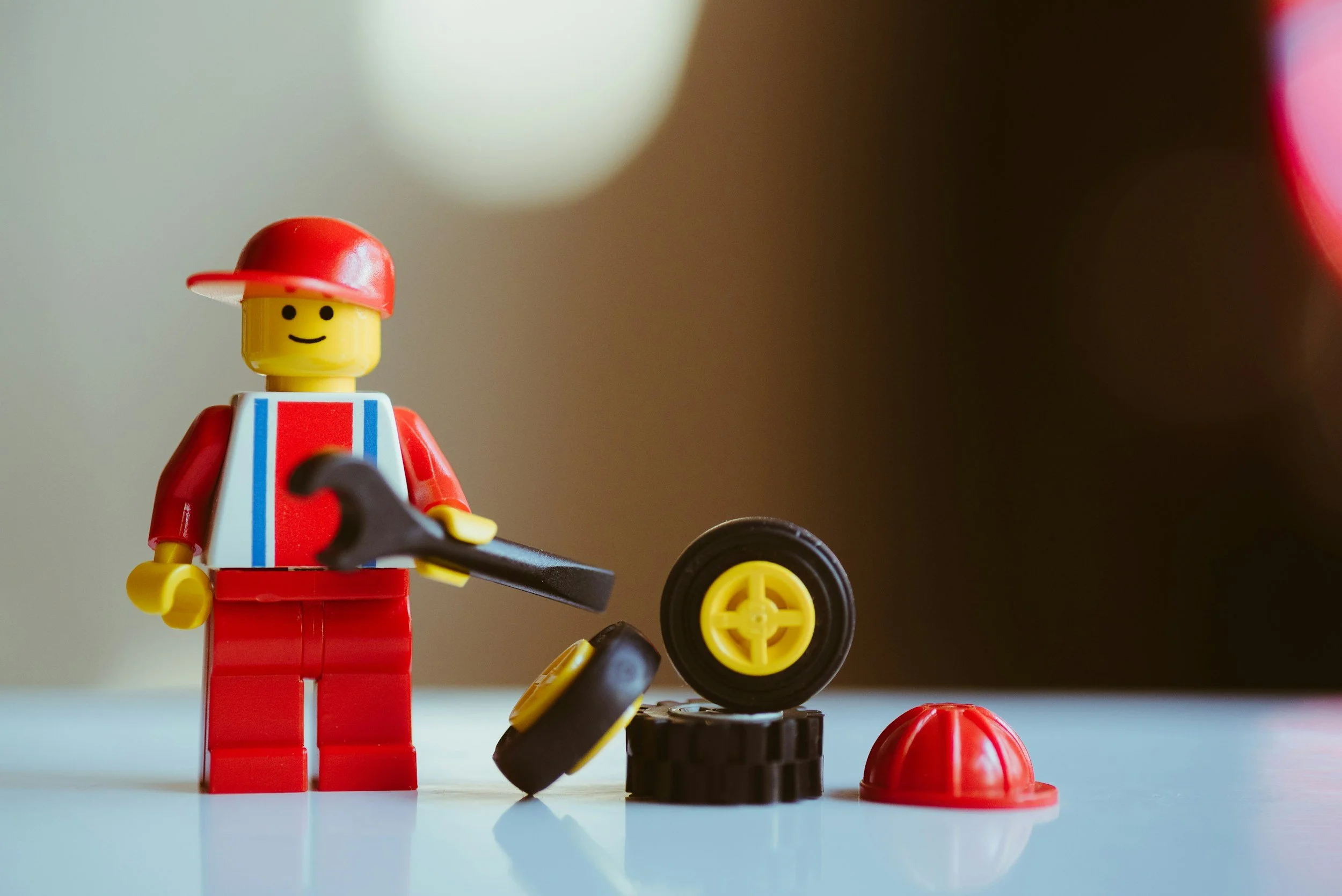 Lego minifigure in red and white outfit with a red cap, holding a wrench, standing next to small black and yellow wheels, with a red Lego hat on the surface.