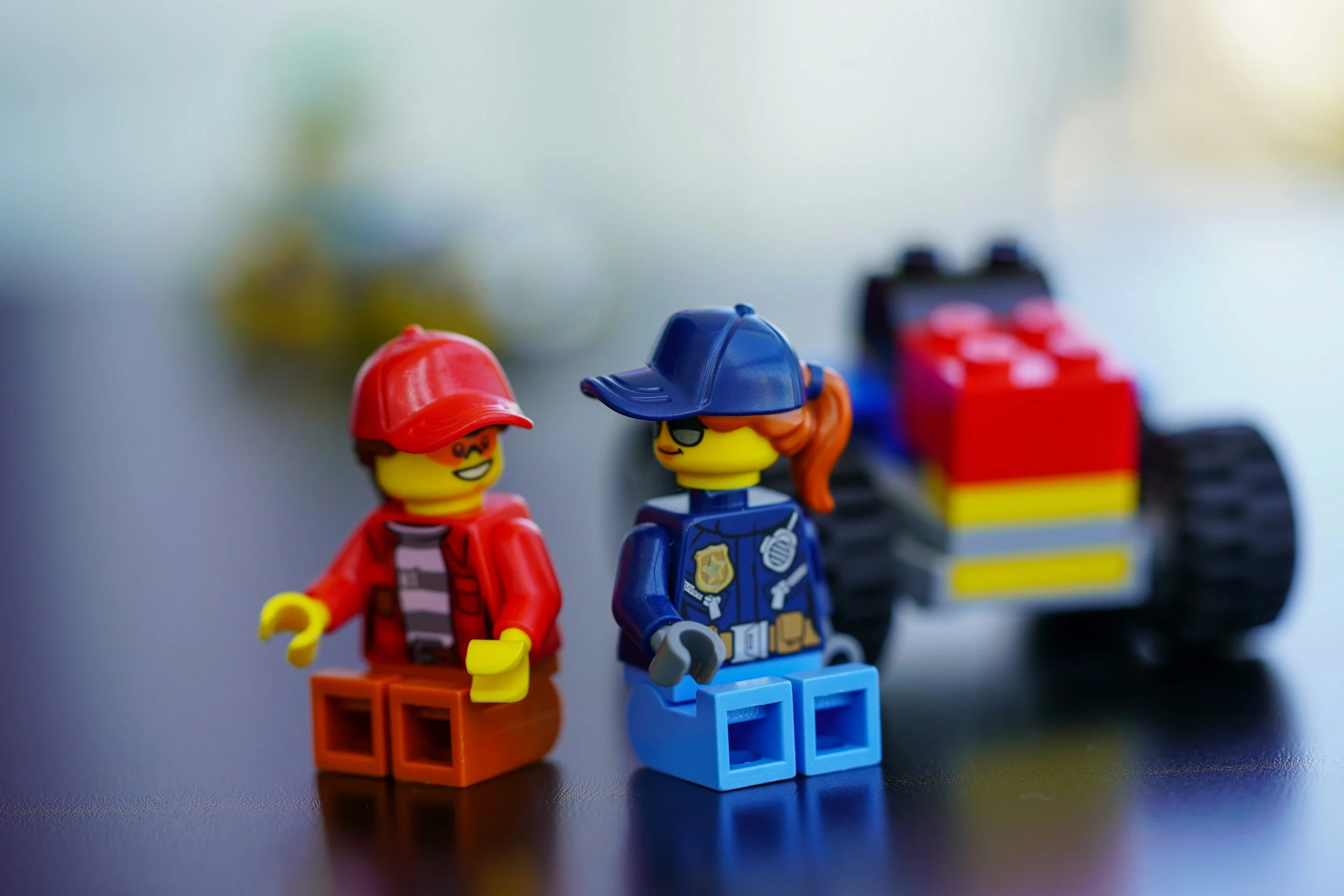 Two LEGO figures, one dressed as a construction worker with a red hat and orange pants, and the other dressed as a police officer with a blue hat and blue uniform, sitting on a black surface with a LEGO vehicle in the background.