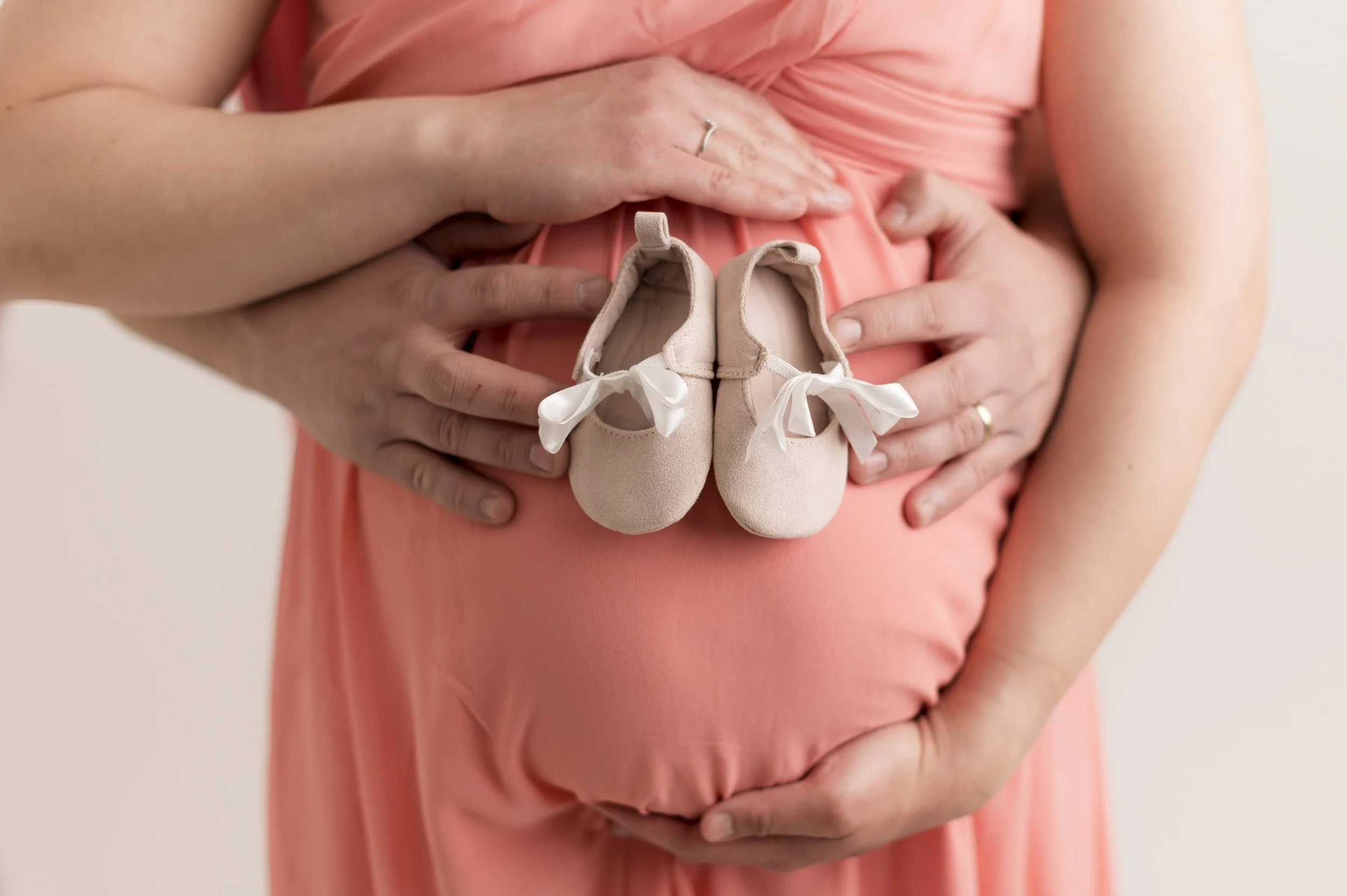 A pregnant woman assisted by her partner, holding baby shoes over her belly. Mom to be needs birth preparation