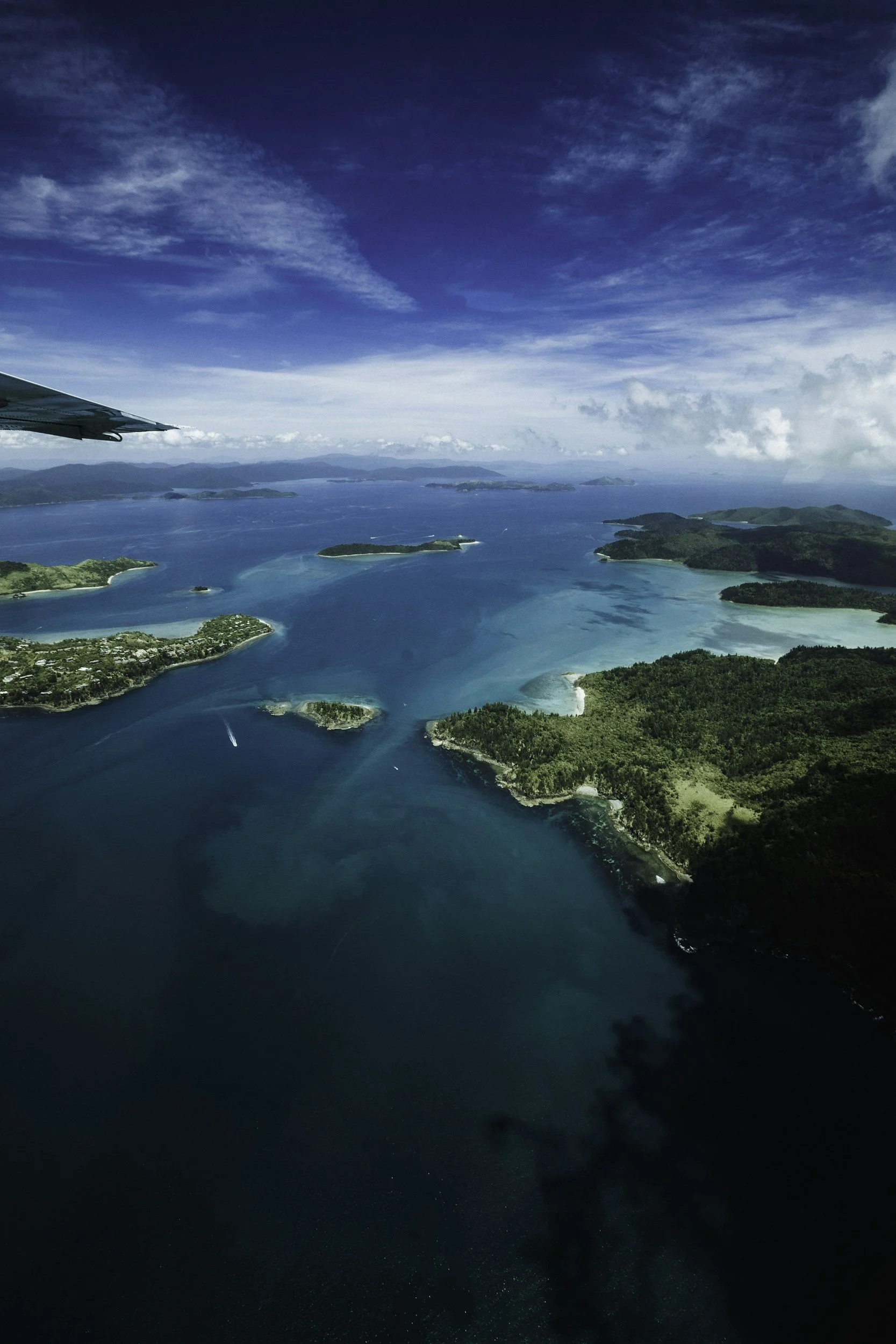 Aerial view of a tropical island surrounded by turquoise waters with lush greenery and small islands, under a partly cloudy sky.