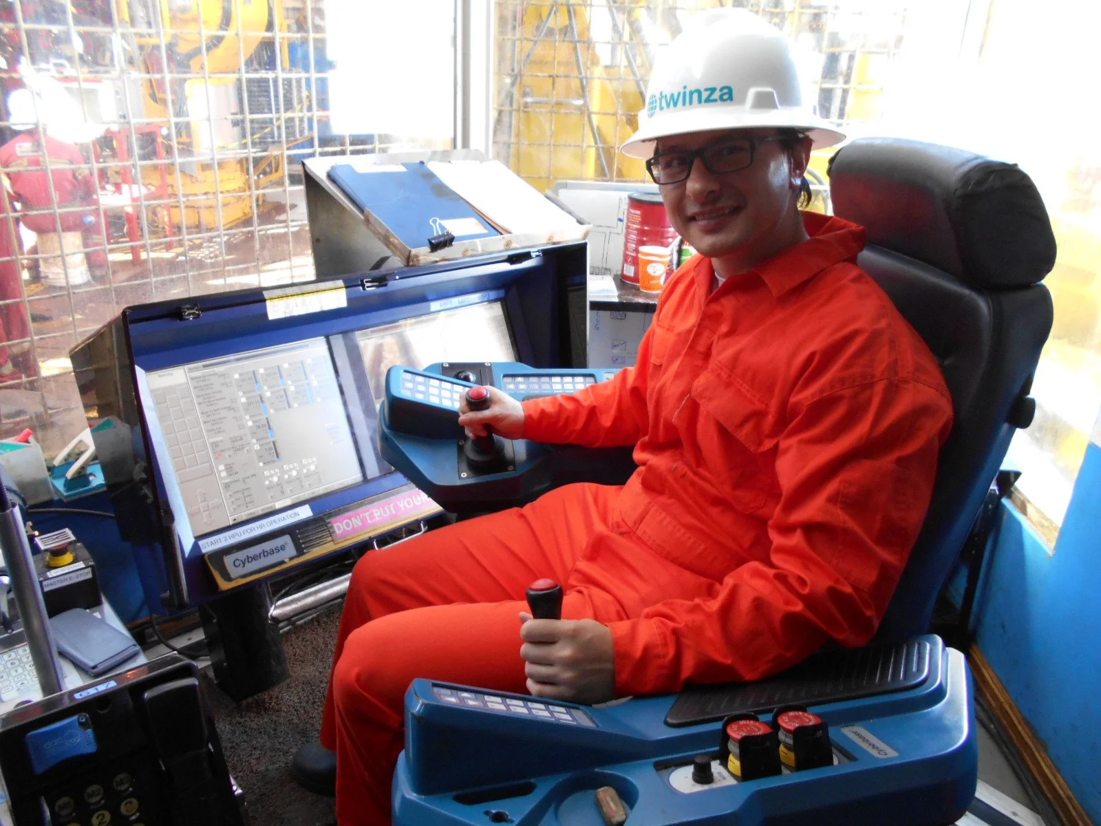 A construction worker in orange coveralls and a white safety helmet is sitting in front of a control panel, smiling at the camera, inside a construction crane cabin.