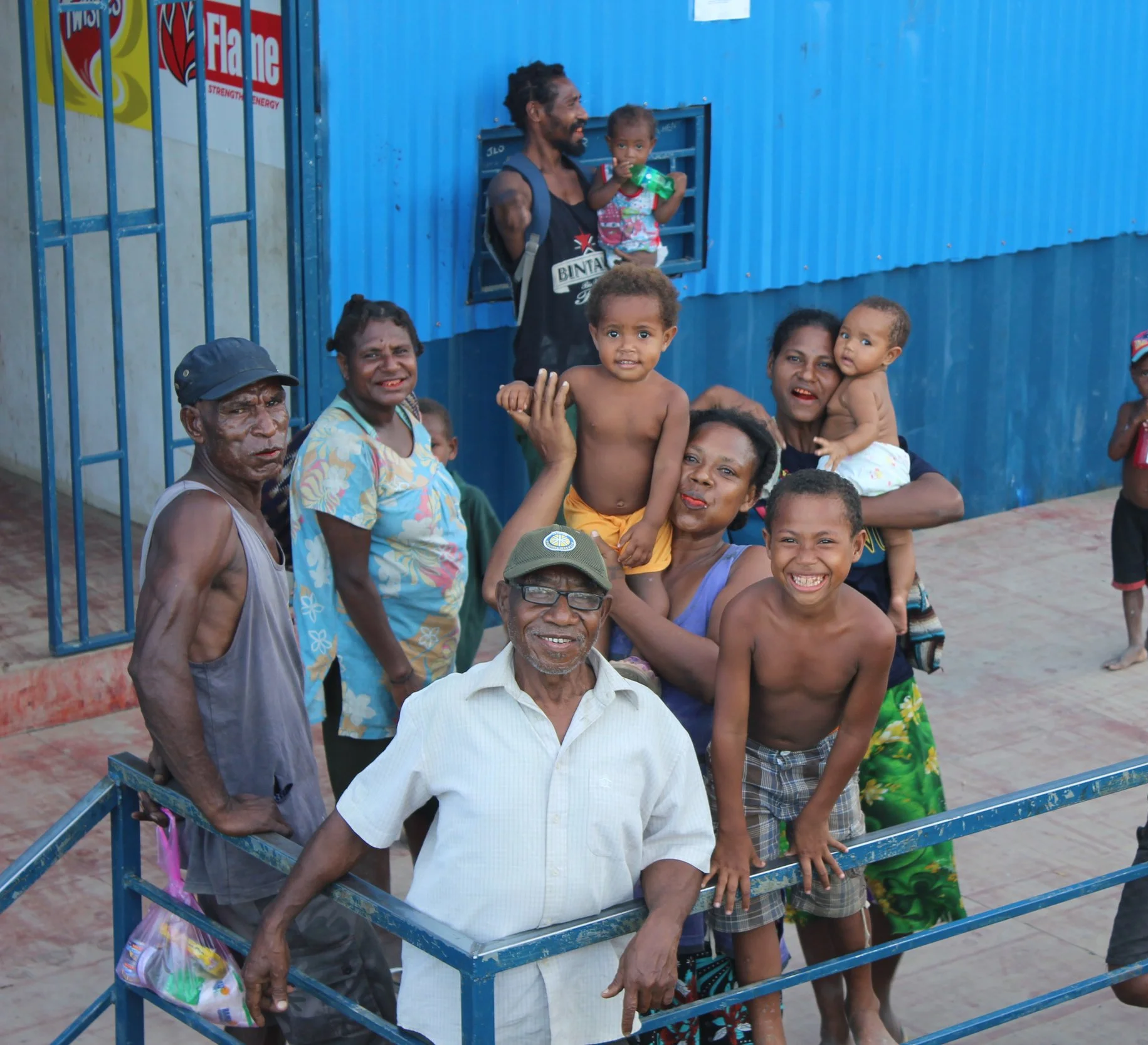 A group of smiling adults and children of various ages gathered outdoors behind a blue metal barrier, with some holding children and others smiling at the camera, in front of a blue building.