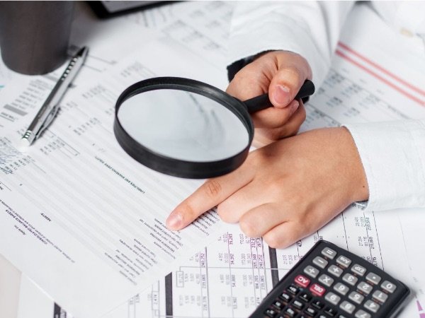 Person using magnifying glass to examine financial documents, with calculator nearby.