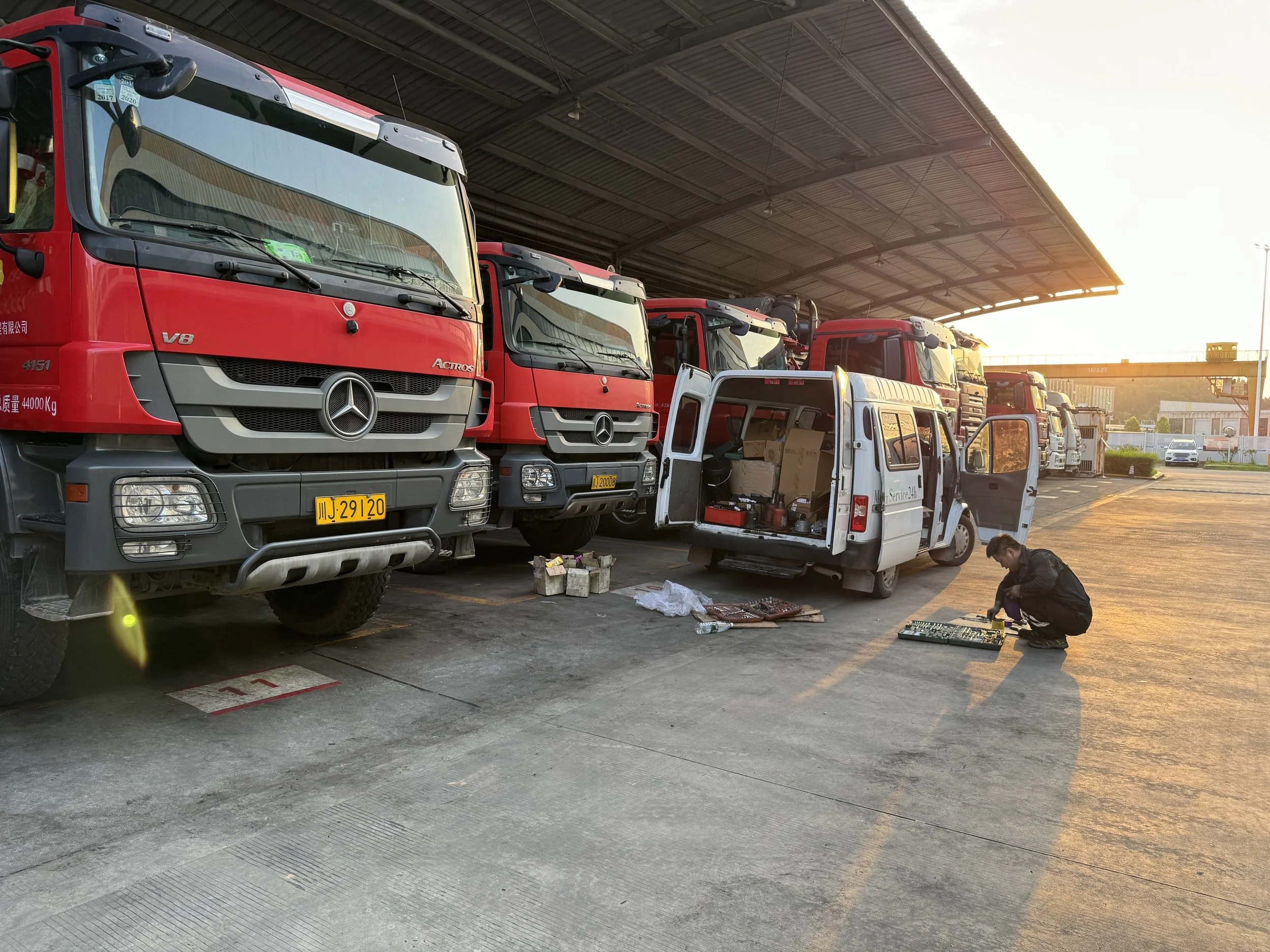 A mechanic working on an electronic circuit board outside at a truck parking lot during sunset, with multiple large red trucks lined up under a canopy.