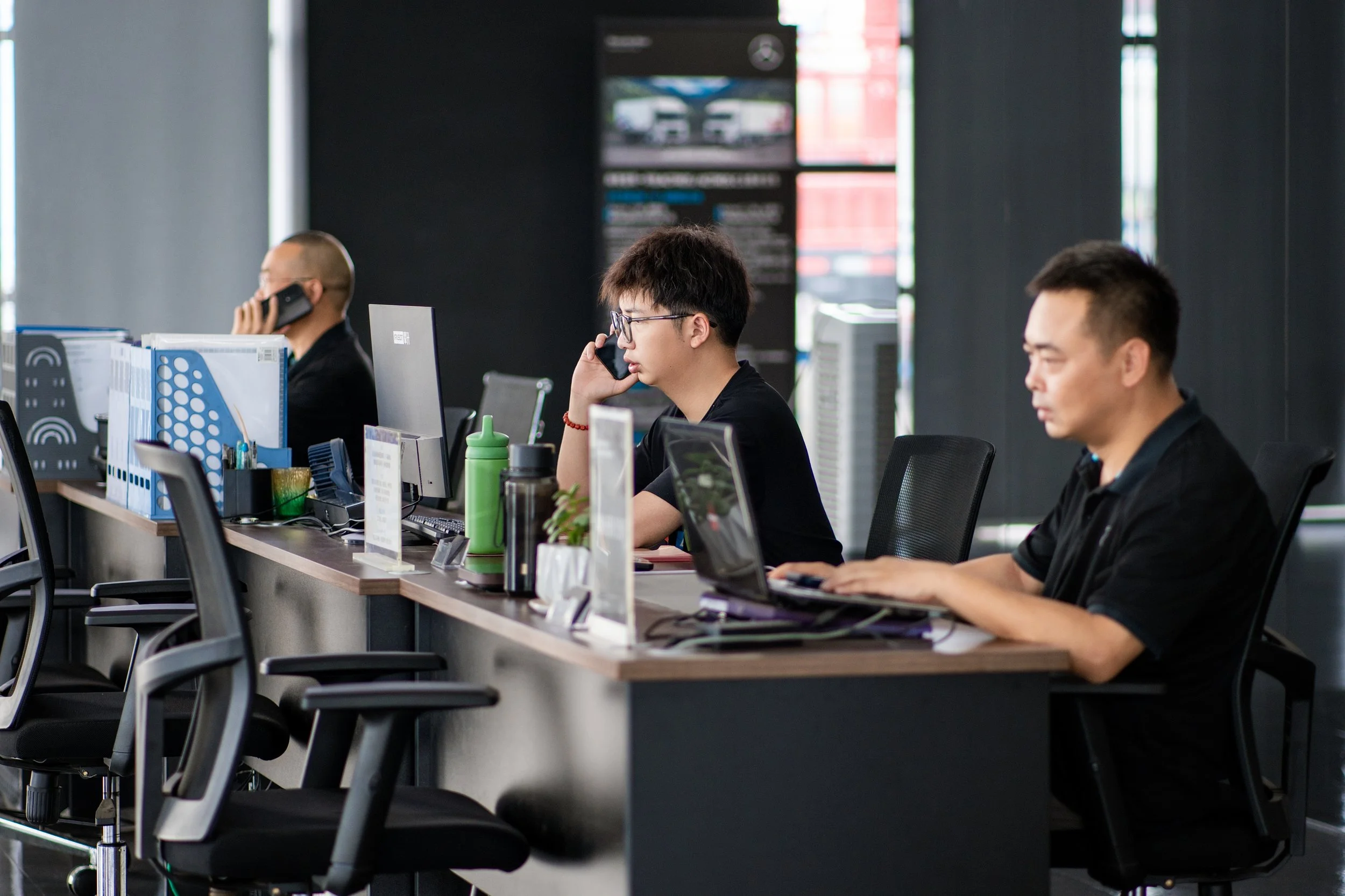 Three men sitting at work desks with computers in an office, using phones and working on laptops.