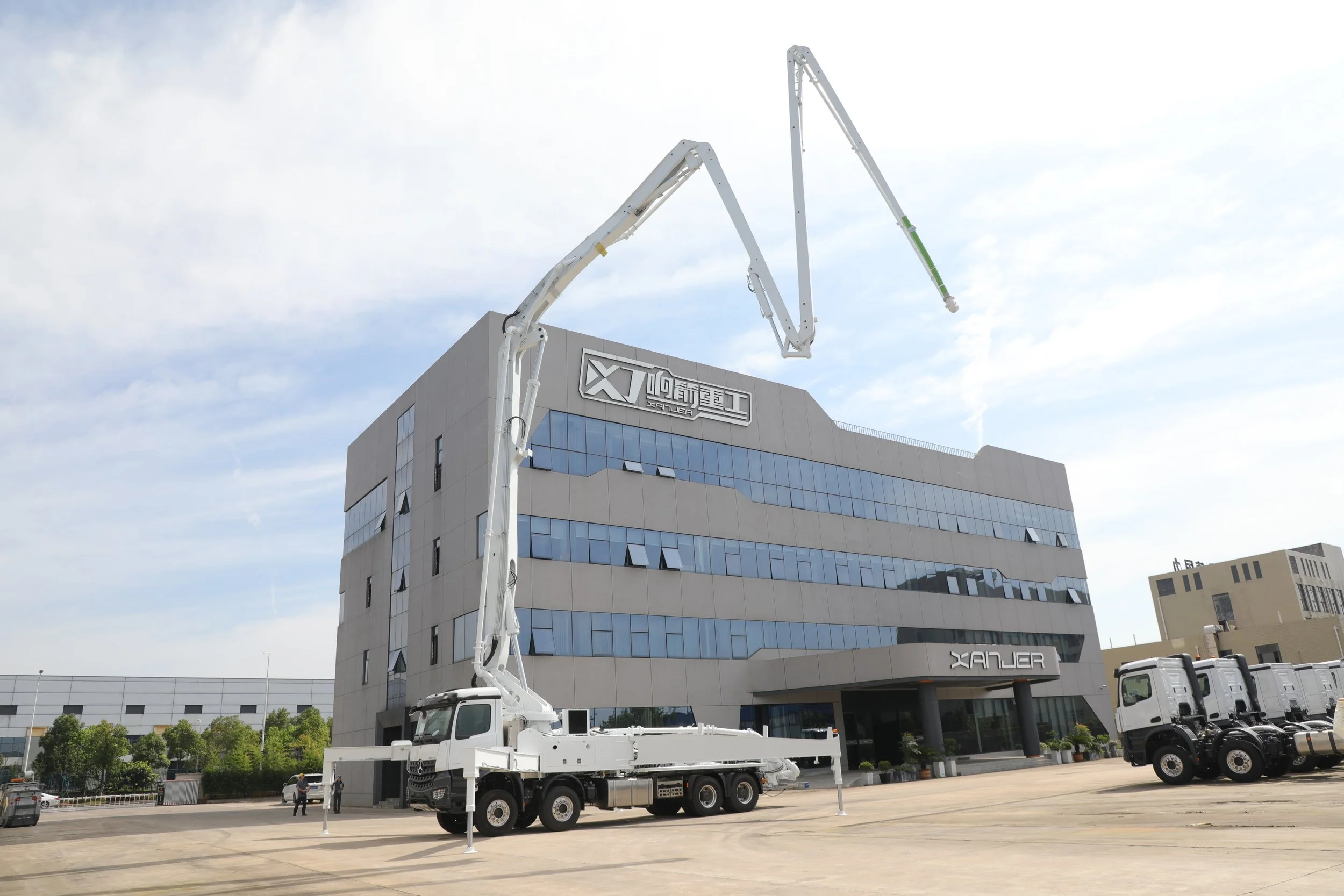 A large white crane truck extending its arm towards the top of a multi-story commercial building with glass windows and signage, parked in an open lot with other trucks nearby.