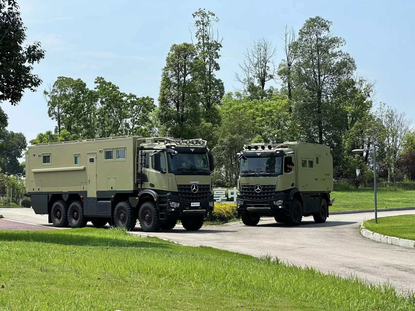 Two military trucks parked on a paved road in a grassy area with trees in the background during daytime.