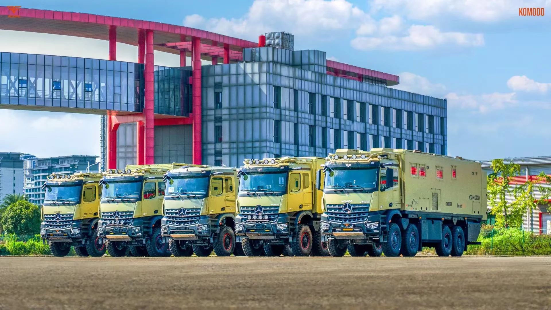 Five yellow military trucks parked in a row outdoors with a modern building in the background under a partly cloudy sky.