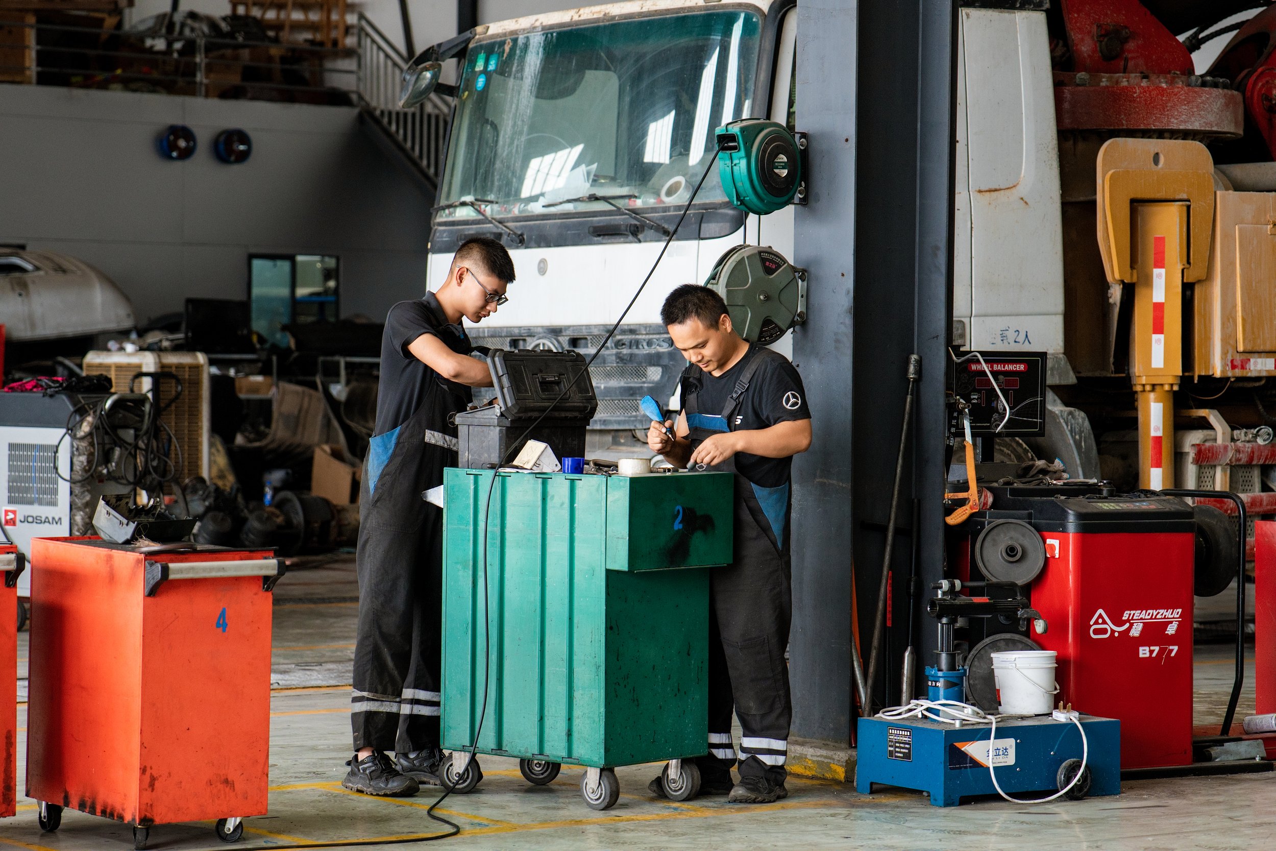 Two male mechanics working together in an industrial garage. They are wearing black overalls and using tools at a green tool cart. The background includes large machinery, a bus, and various equipment.