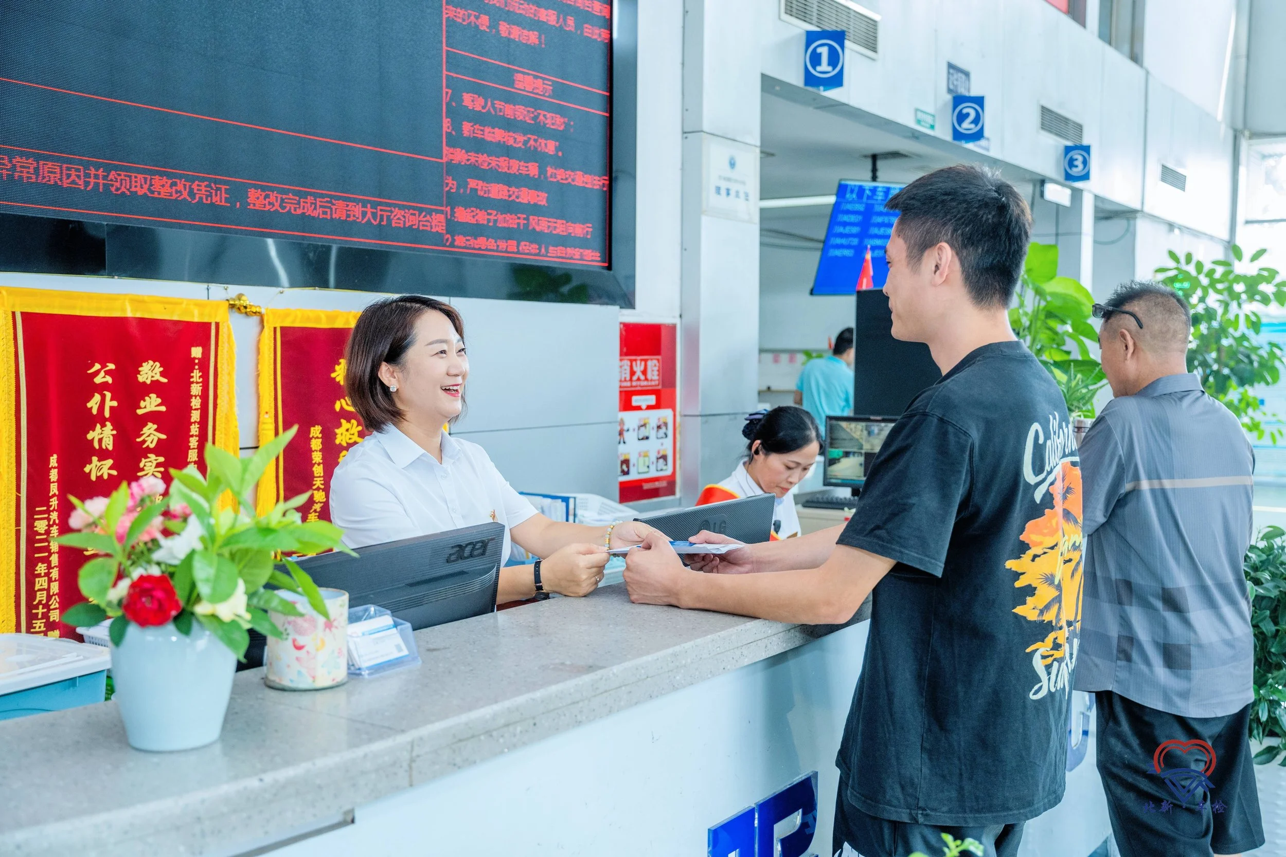 A customer service counter at a government office with a woman employee smiling and handing a document to a young man. There are other staff members working, a large digital display with red text, and decorative banners behind the counter. Potted pla