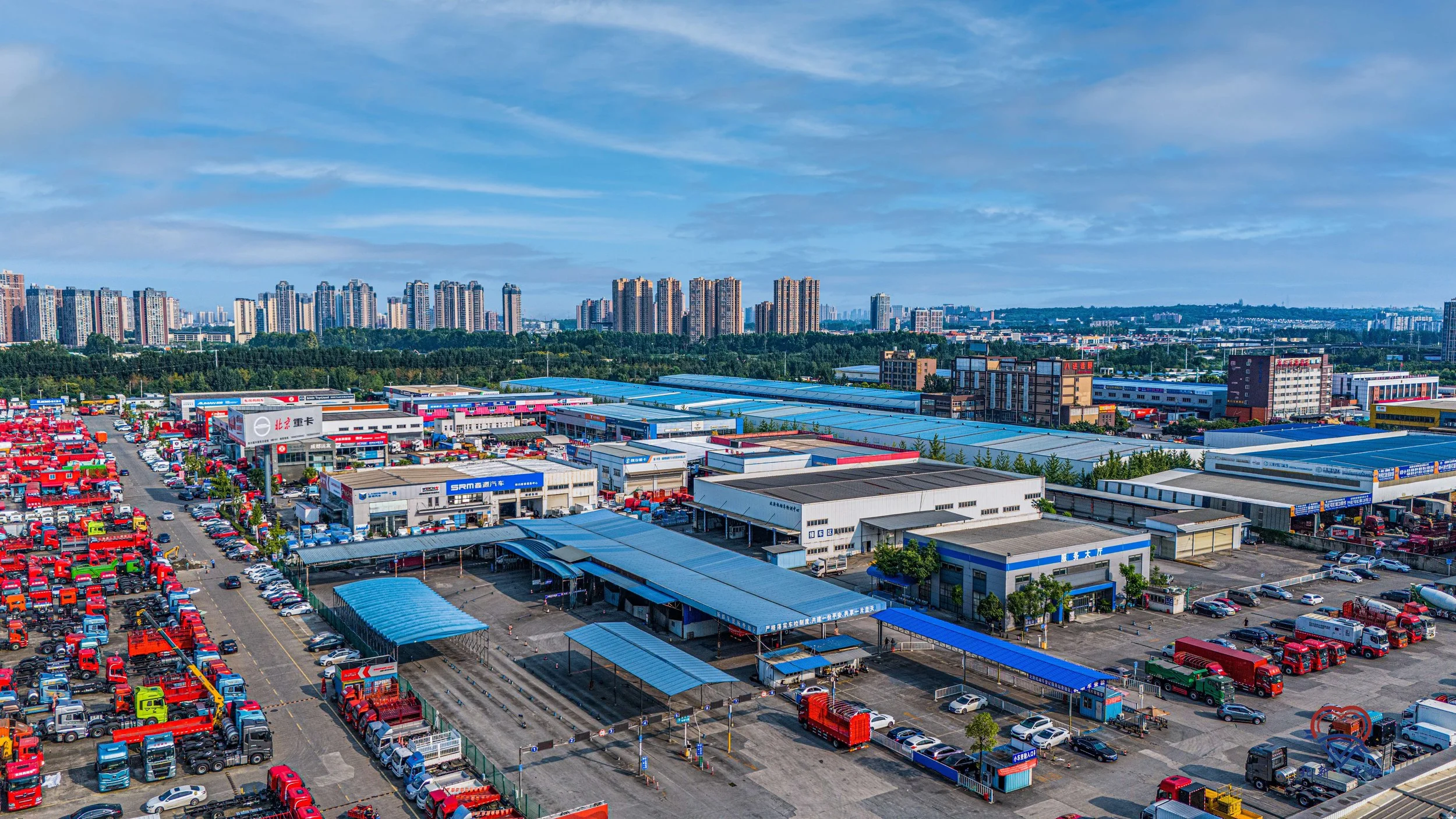 A large industrial warehouse and truck yard with many trucks parked, surrounded by a cityscape with tall buildings and a blue sky with some clouds.