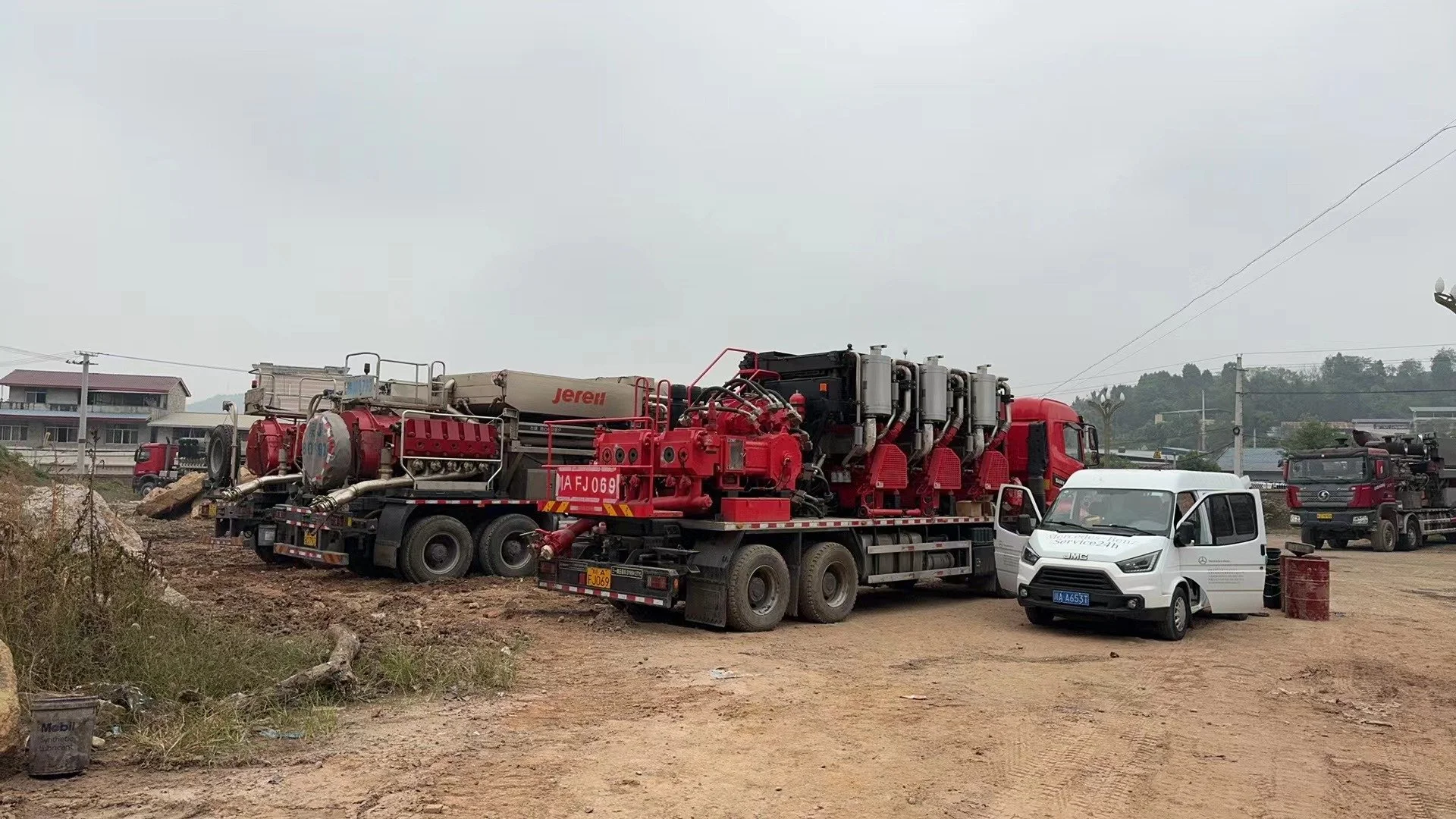 Several large fire trucks and equipment vehicles parked on a dirt lot with gray overcast sky, some small buildings and power lines in background.