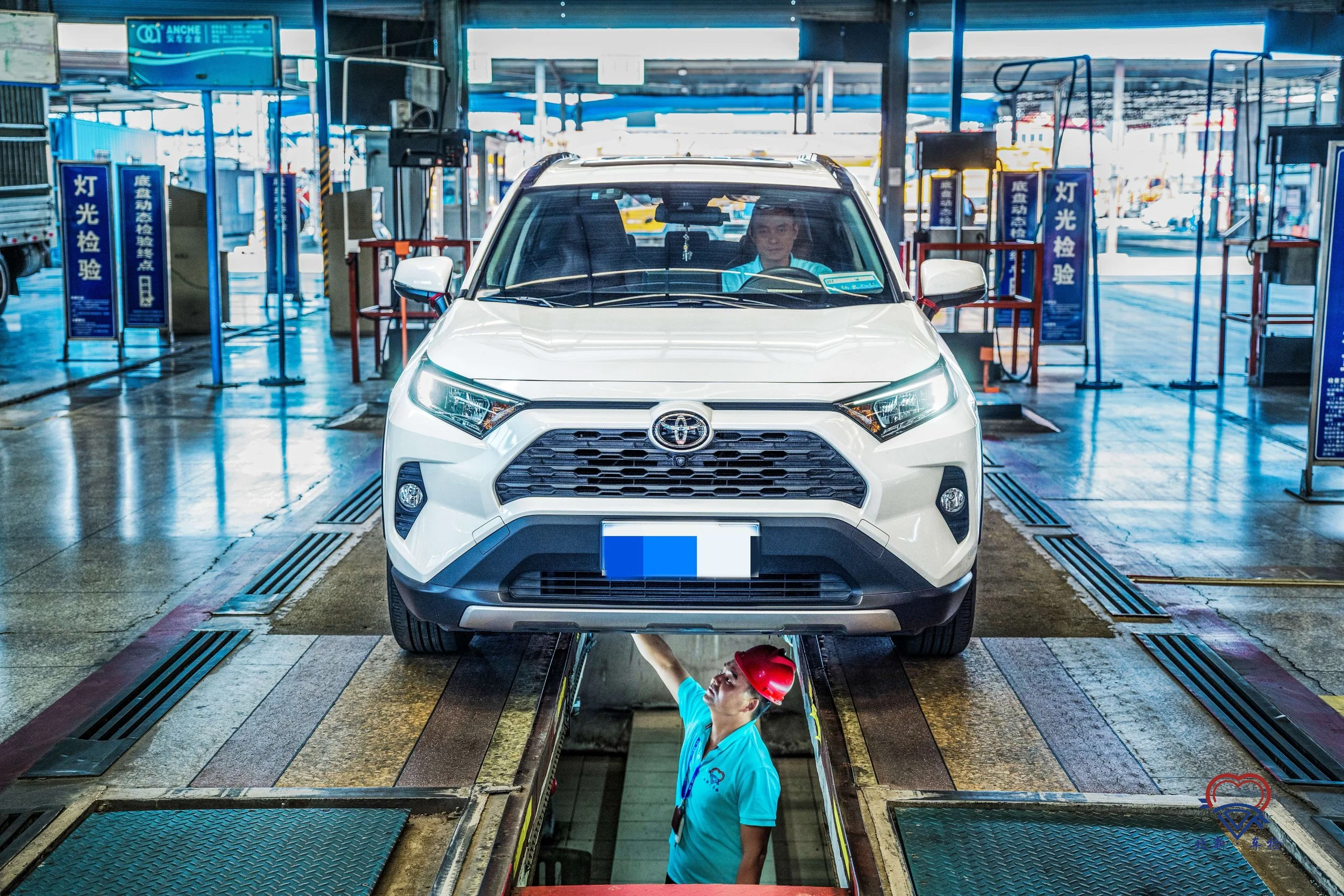 A white Toyota SUV is on a vehicle testing or inspection platform inside a service center, with a technician underneath reaching up to work on it, wearing a red safety helmet and blue uniform.