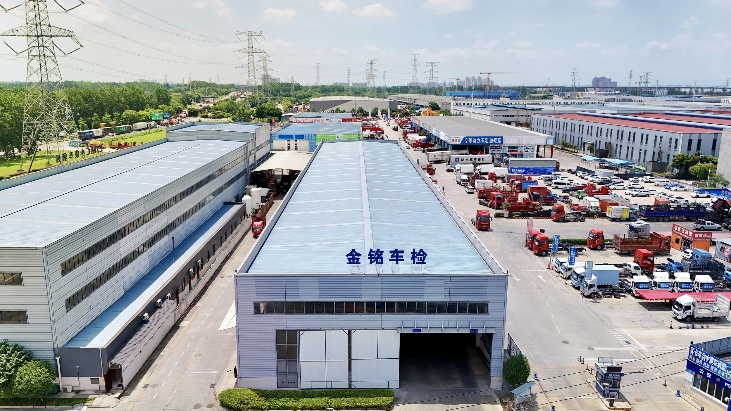 Aerial view of a large industrial truck inspection yard with multiple warehouse buildings, parked trucks, and cranes, with power lines and trees in the background.