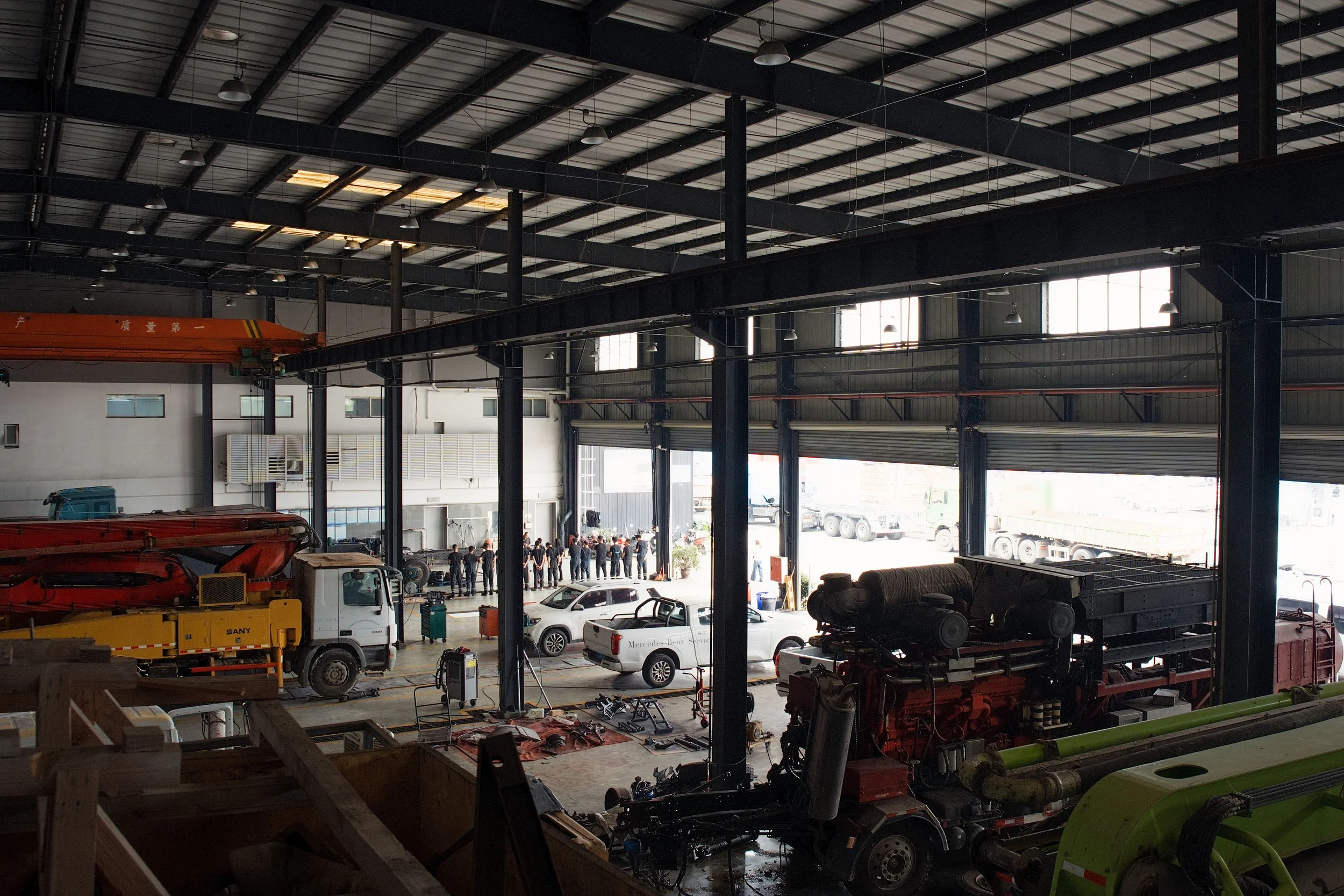 Inside a large industrial warehouse with vehicles and machinery, with a line of people gathered near the open entrance.