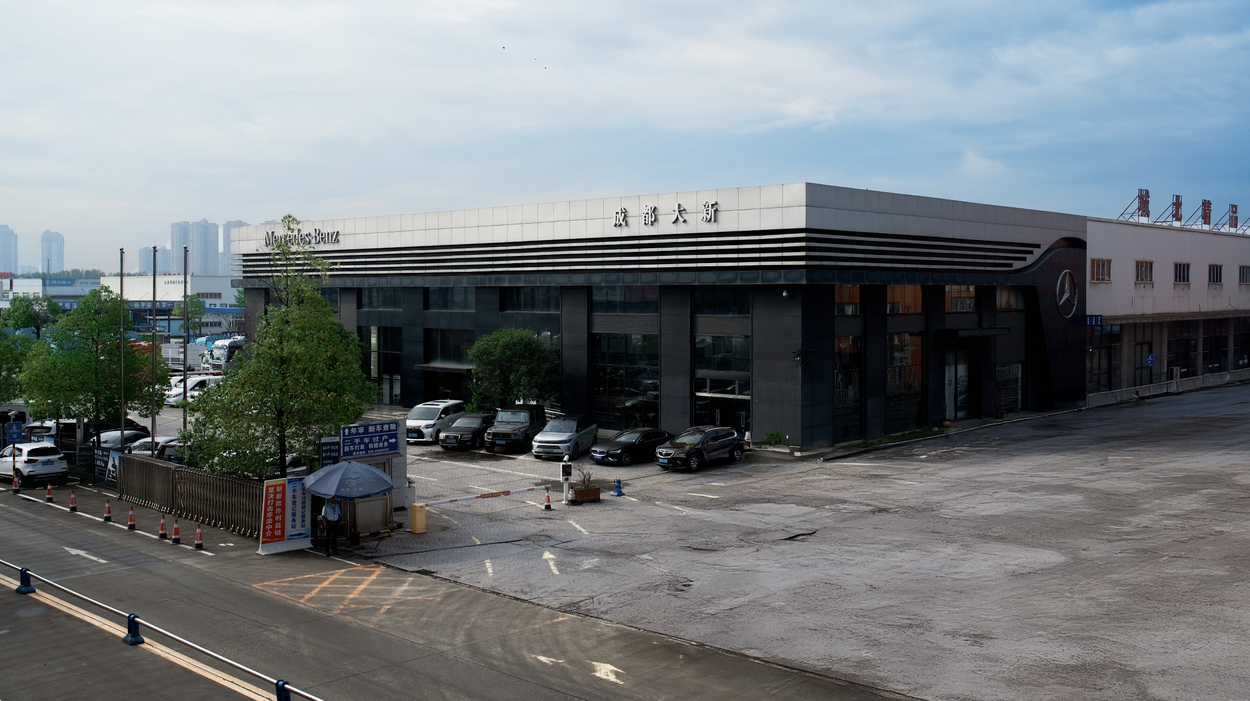 A Mercedes Benz dealership building with parking lot in front, some cars parked, and trees around. The building has signage in Chinese characters and the Mercedes Benz logo.