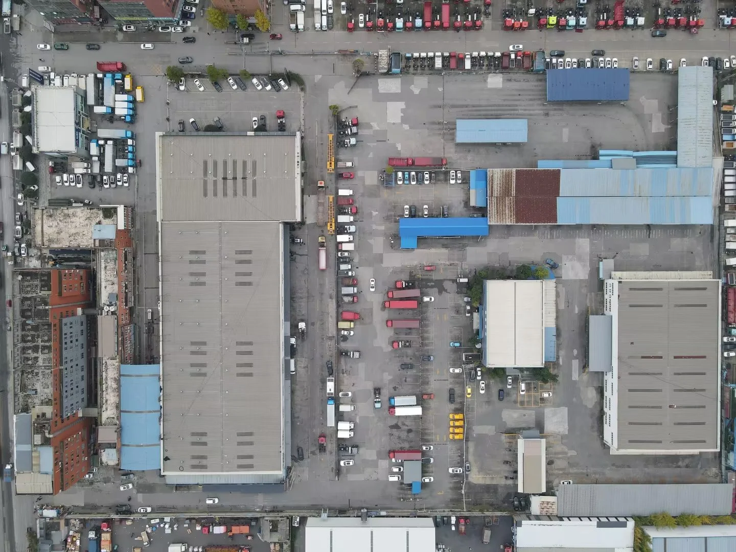 An aerial view of a parking lot filled with cars, trucks, and buses, surrounding large industrial or commercial buildings with grey rooftops, some with blue roofs and some with rusting or weathered sections. The parking lots have vehicles of various