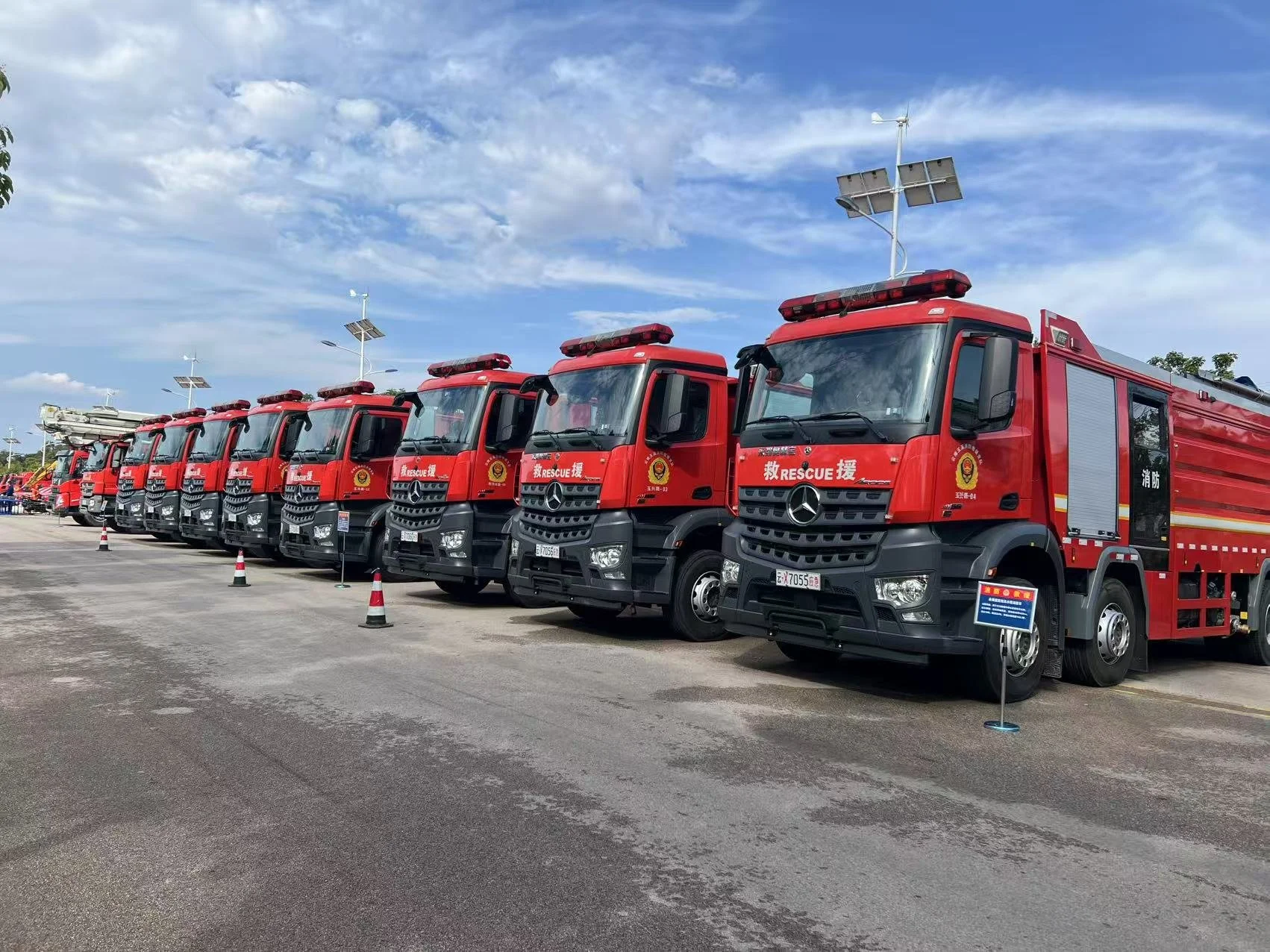 Line of six red fire trucks parked side by side on a paved lot with a blue sky and white clouds above, each with solar panel antennas on the roof.