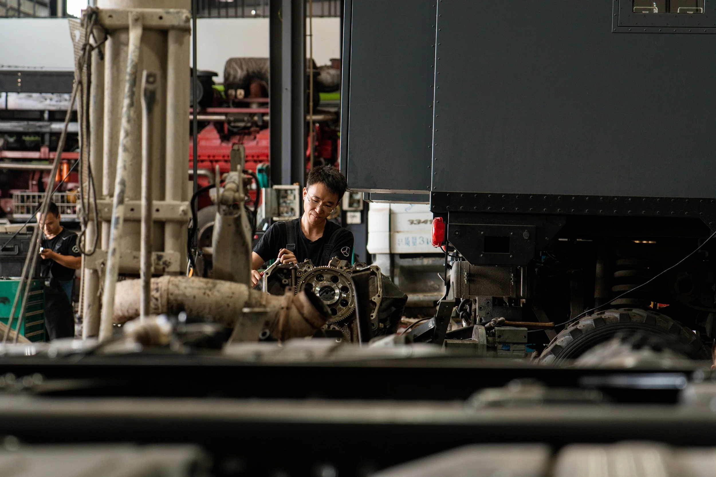 Two workers in a machine repair shop, one in the foreground inspecting or working on a mechanical component, and another worker in the background, both wearing black shirts.