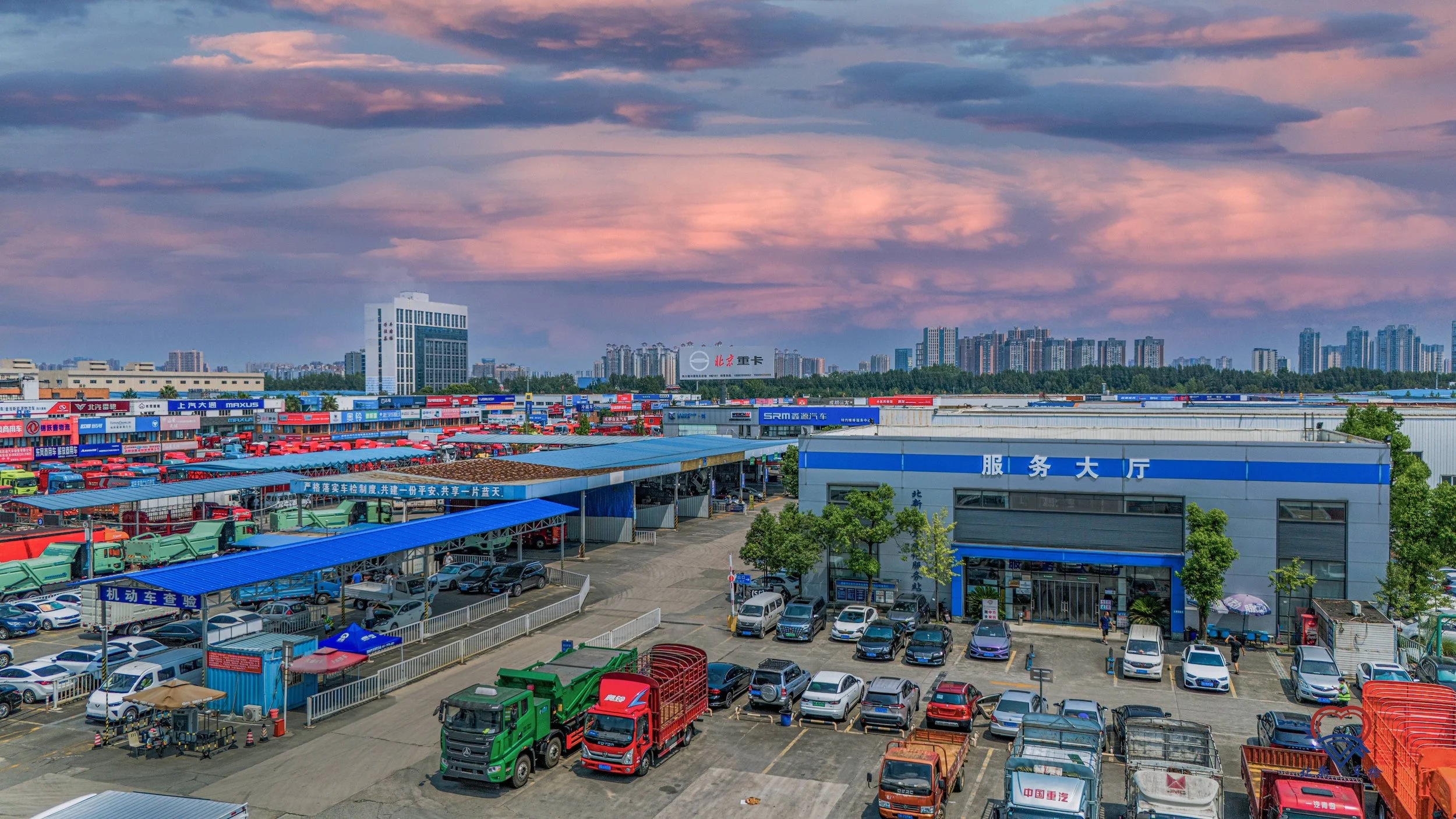 A busy parking lot with various cars and trucks in front of a large building with Chinese characters and a blue and gray color scheme. In the background, there's a wide commercial area with many shops and buildings, and a distant city skyline under a partly cloudy sky at sunset.