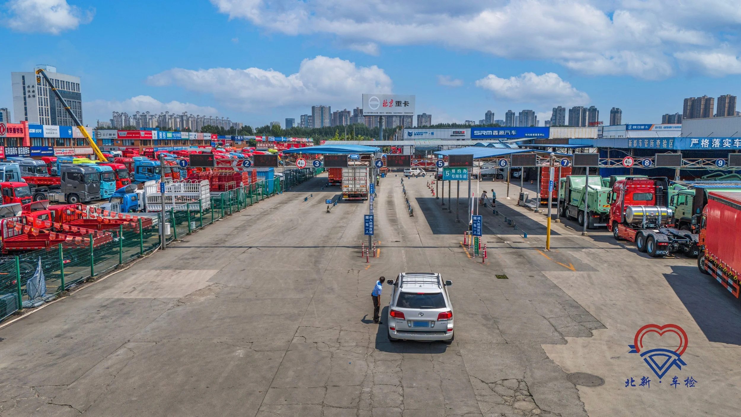 A truck inspection yard with various trucks parked in designated lanes, some with blue, red, green, and gray colors. A person in a blue shirt stands near a silver SUV, speaking to someone inside. There are signs and barriers guiding the inspection pr