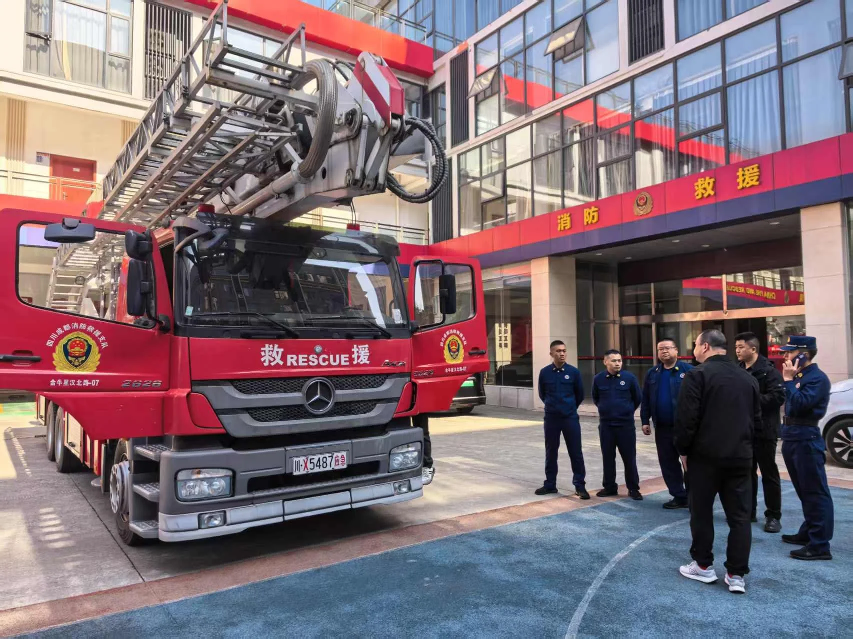 Group of firefighters and officials standing outside a fire rescue truck in front of a building with Chinese characters on the sign.