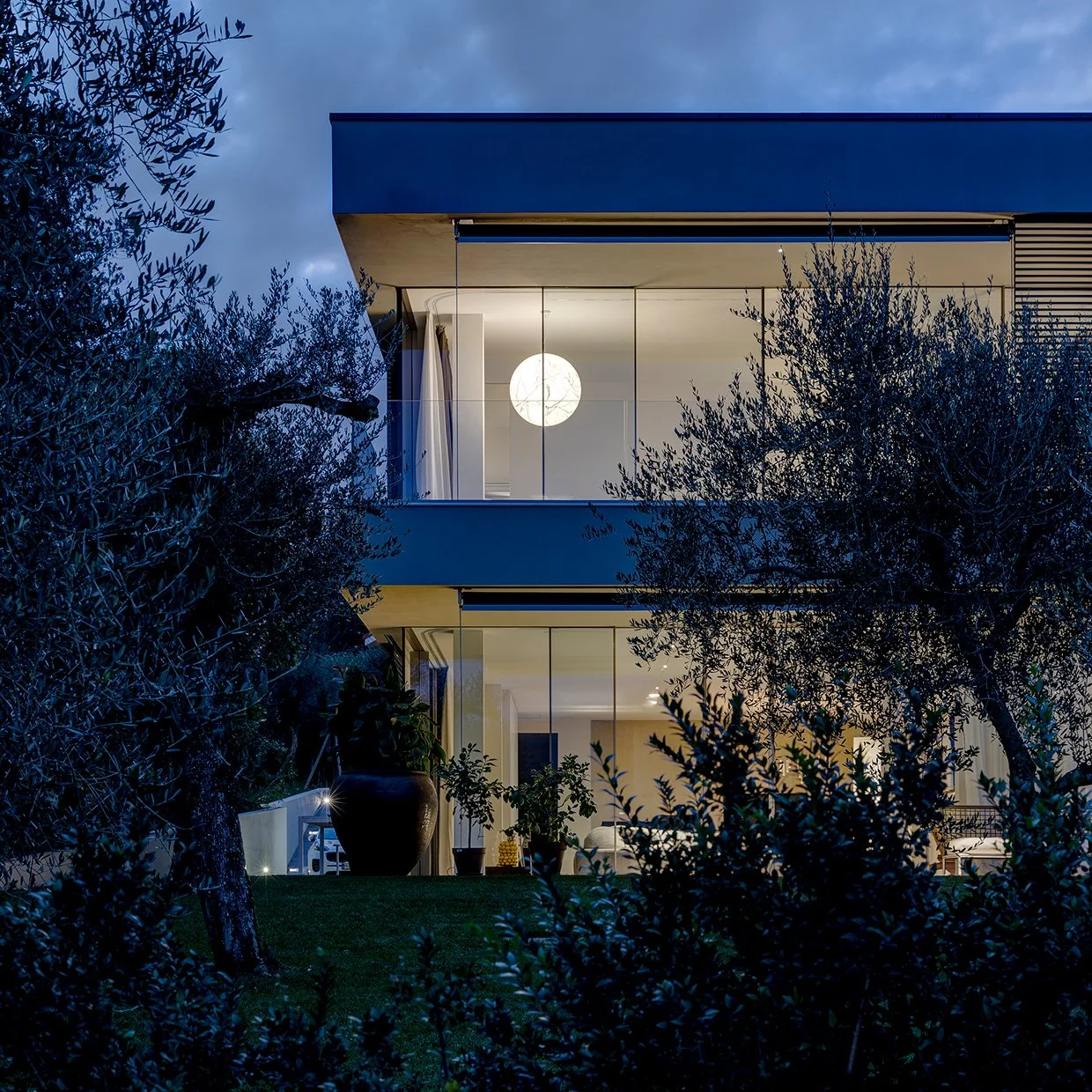 Nighttime exterior view of a modern multi-story house with large glass windows, illuminated interior, surrounding trees, and a glowing spherical moon visible through the upper window.