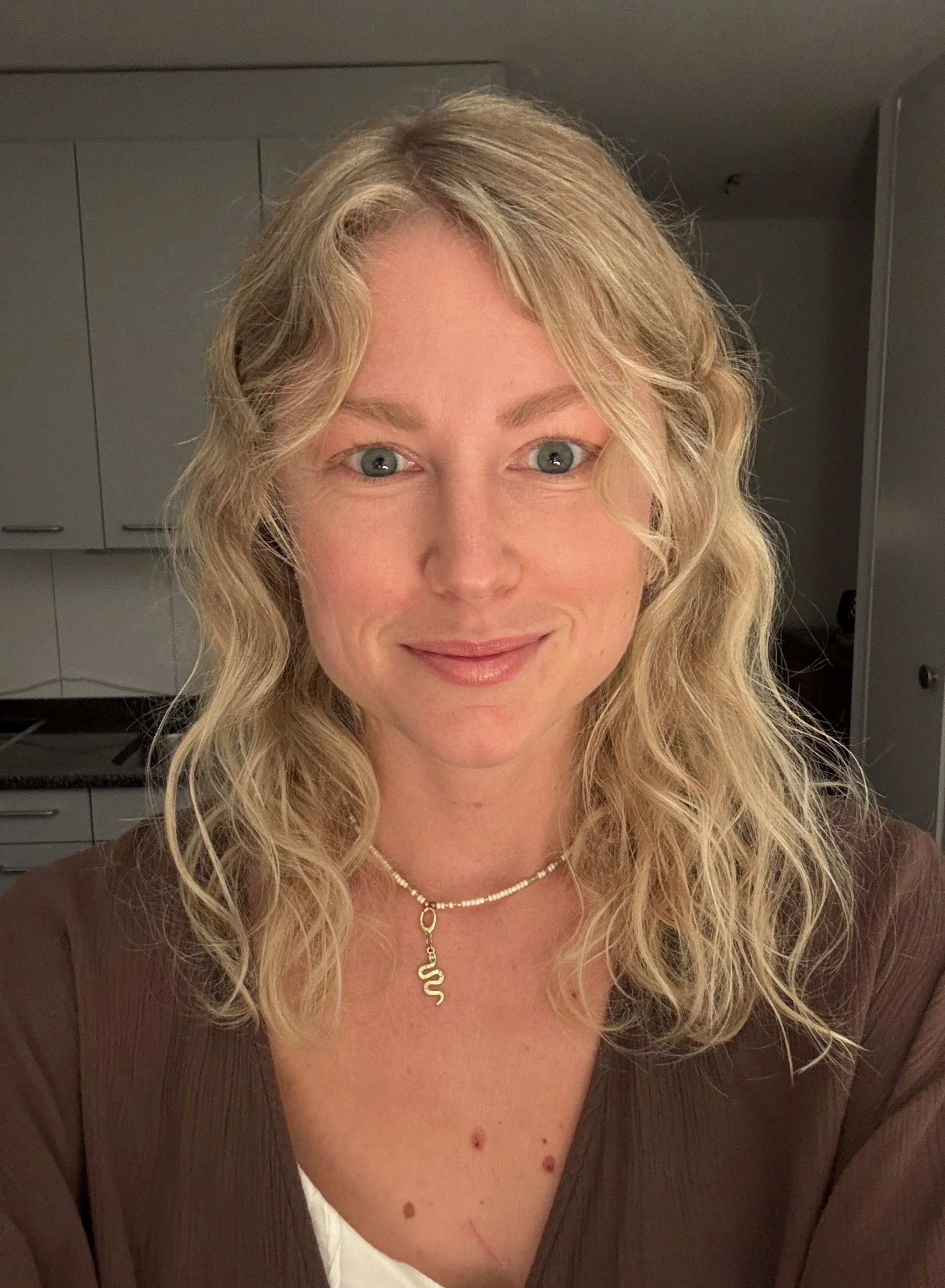 Ein Porträtfoto einer Frau mit blonden, welligen Haaren, blauen Augen und einem freundlichen Lächeln, die eine Halskette trägt, in einer Küche.