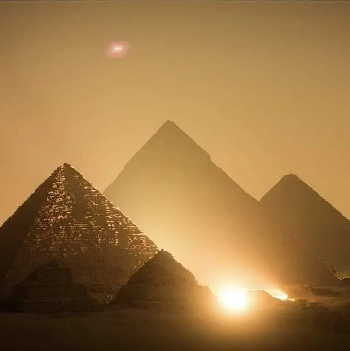 The image shows the pyramids of Egypt at night with a bright light source illuminating them from the bottom, creating an ethereal glow and a slightly cloudy atmosphere.