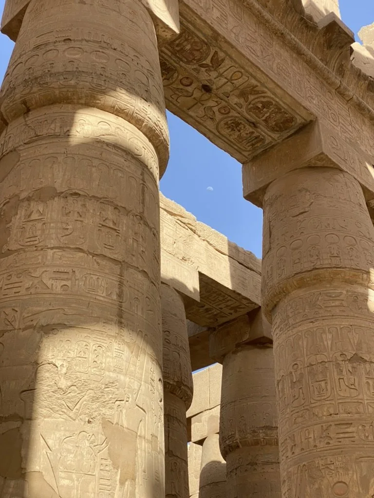 Ancient Egyptian stone columns with hieroglyphics and carvings, part of an archaeological site or temple, under a clear blue sky.