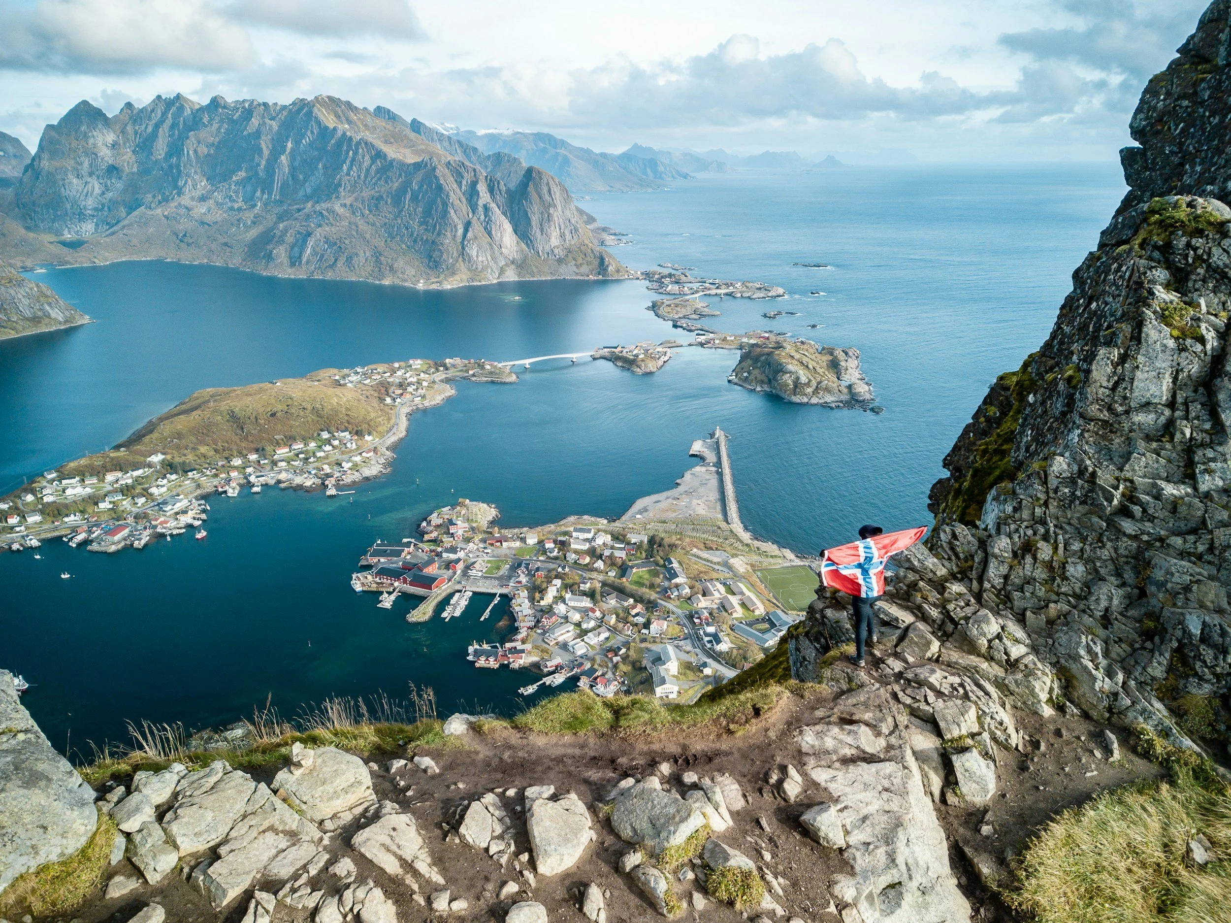 Person on rocky cliff holding a Norwegian flag, overlooking a fjord with a small town and mountains in the background.