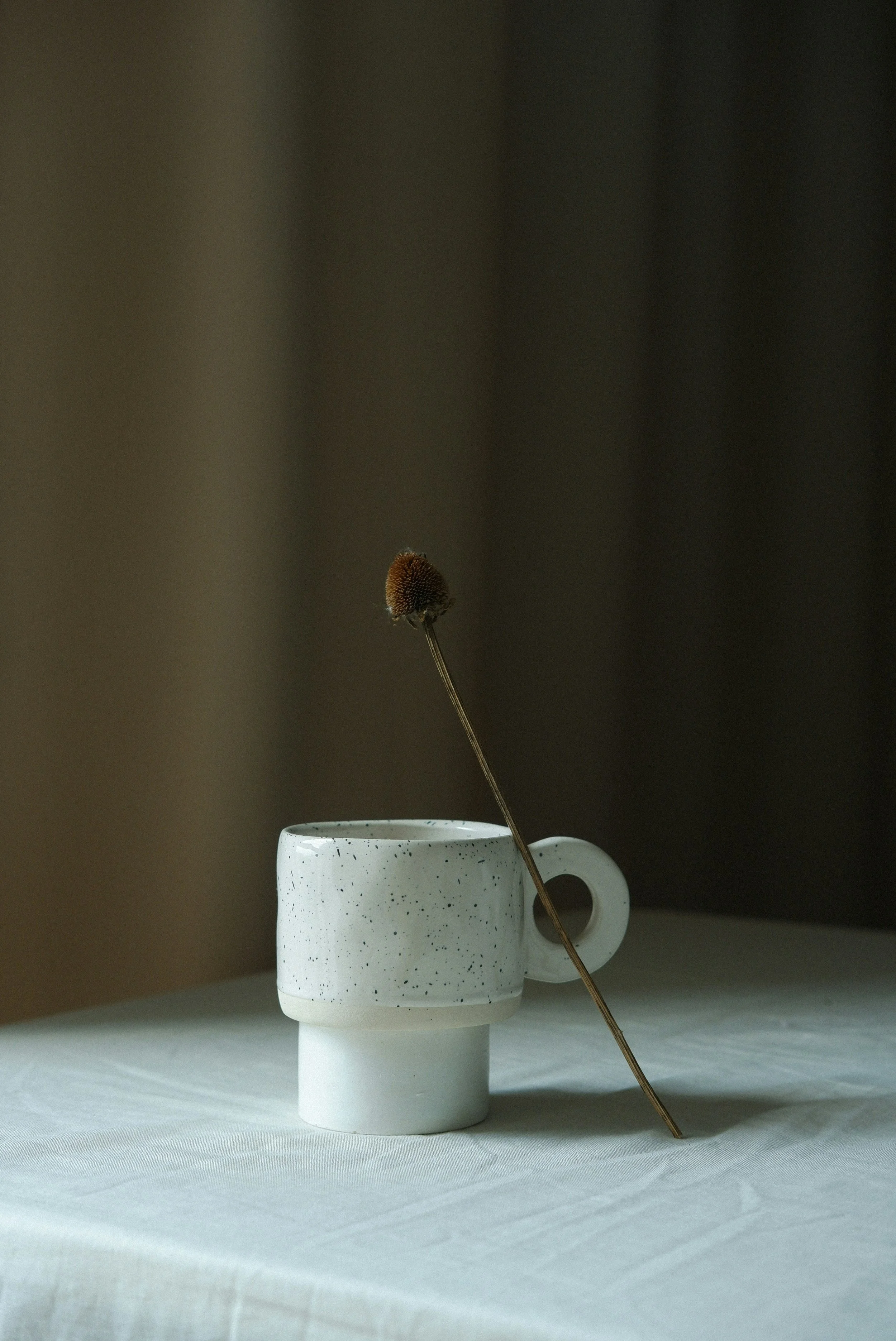 A speckled white ceramic cup with a circular handle, placed on a white tablecloth, with a dried flower resting inside it.