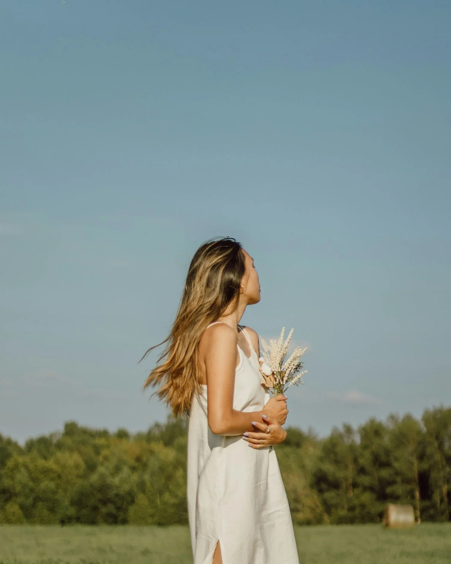 A woman in a white dress holding a small bouquet of dried flowers, looking to her right outdoors with a field and trees in the background under a clear blue sky.