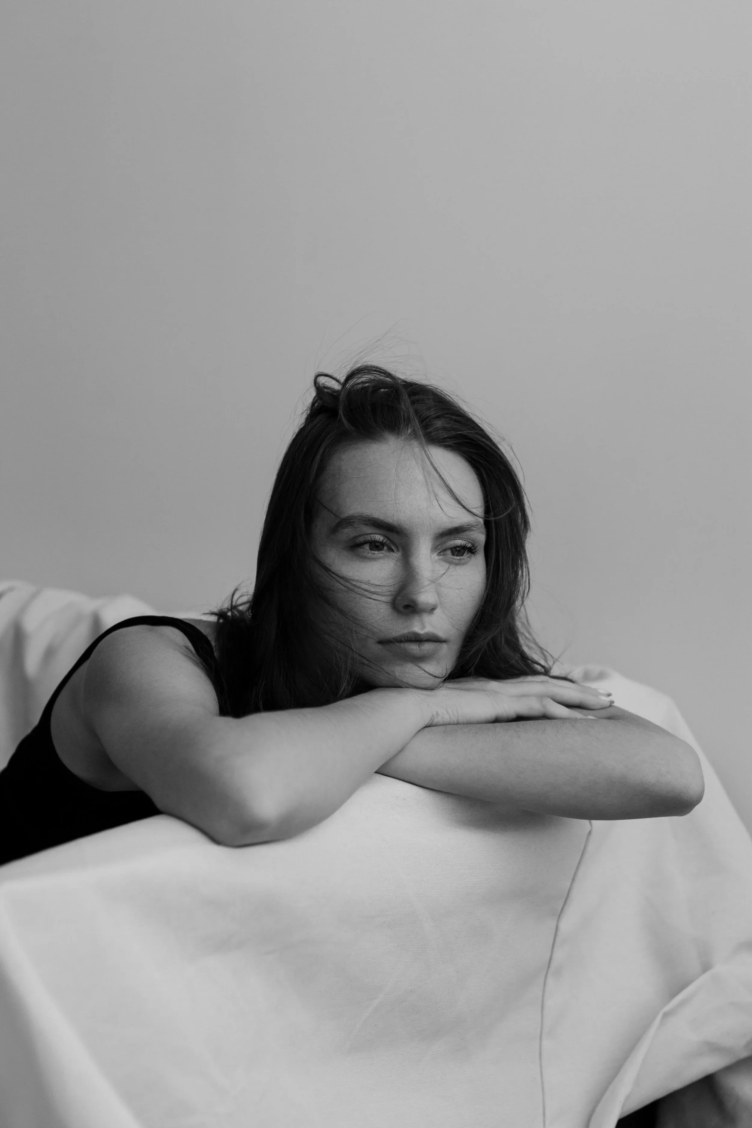 Black-and-white photo of a young woman with dark hair resting her head and arms on a piece of furniture, gazing thoughtfully into the distance.