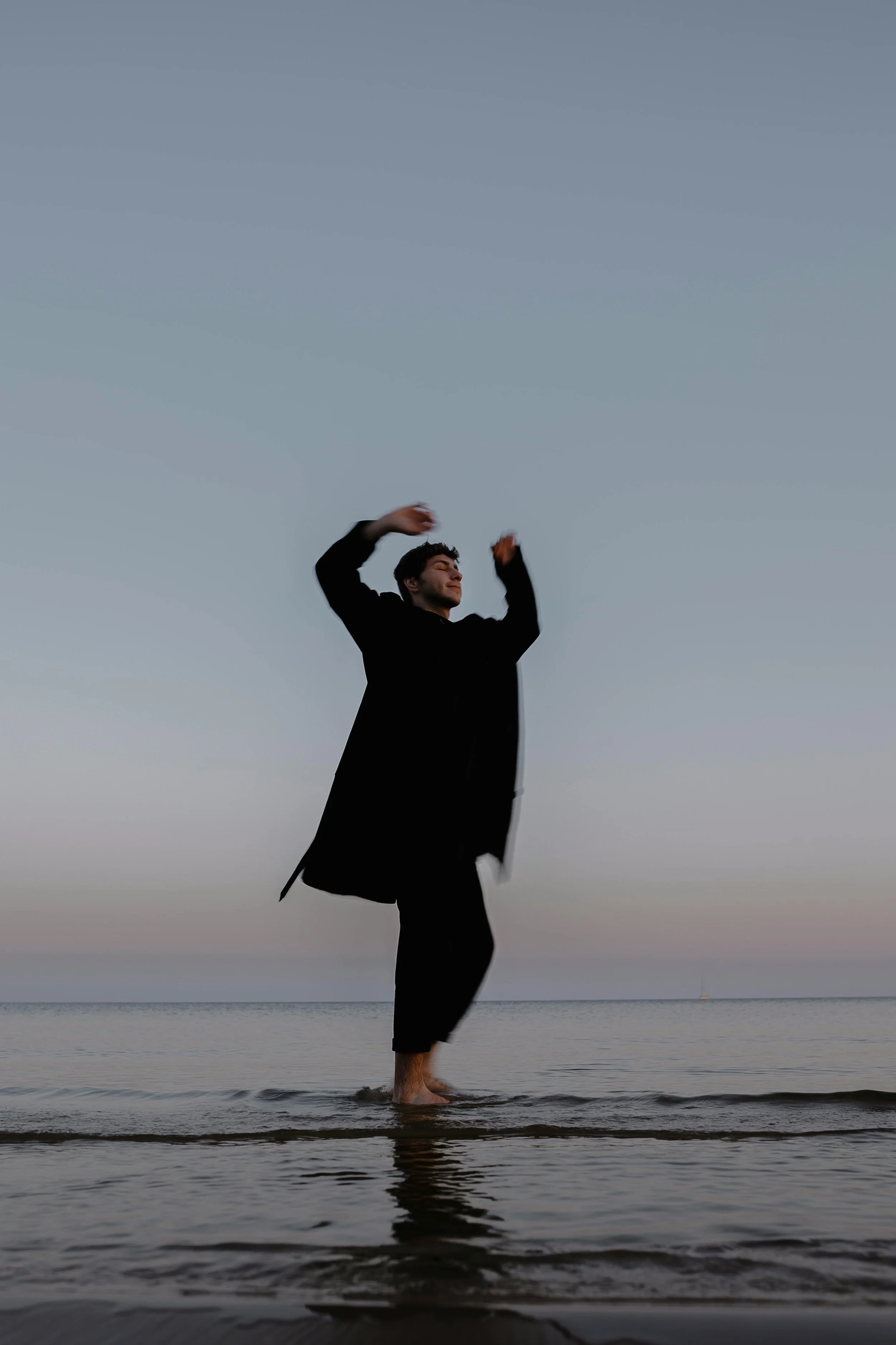 A young man in black clothing standing in shallow water at the beach during sunset, with arms raised and eyes closed, appearing to enjoy the moment.