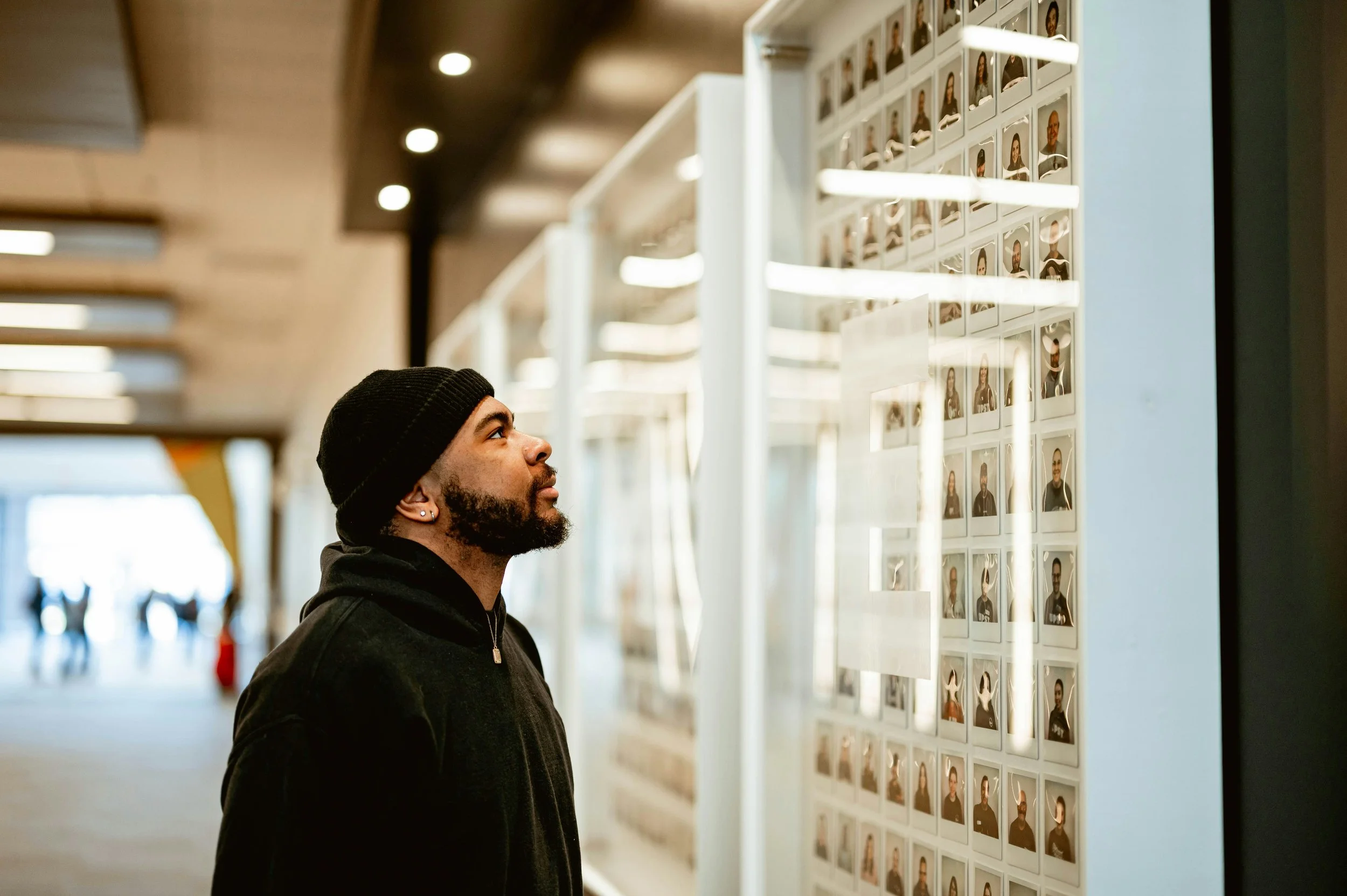 A man with a beard, black beanie, and black jacket looking at a photo display in an indoor public space.