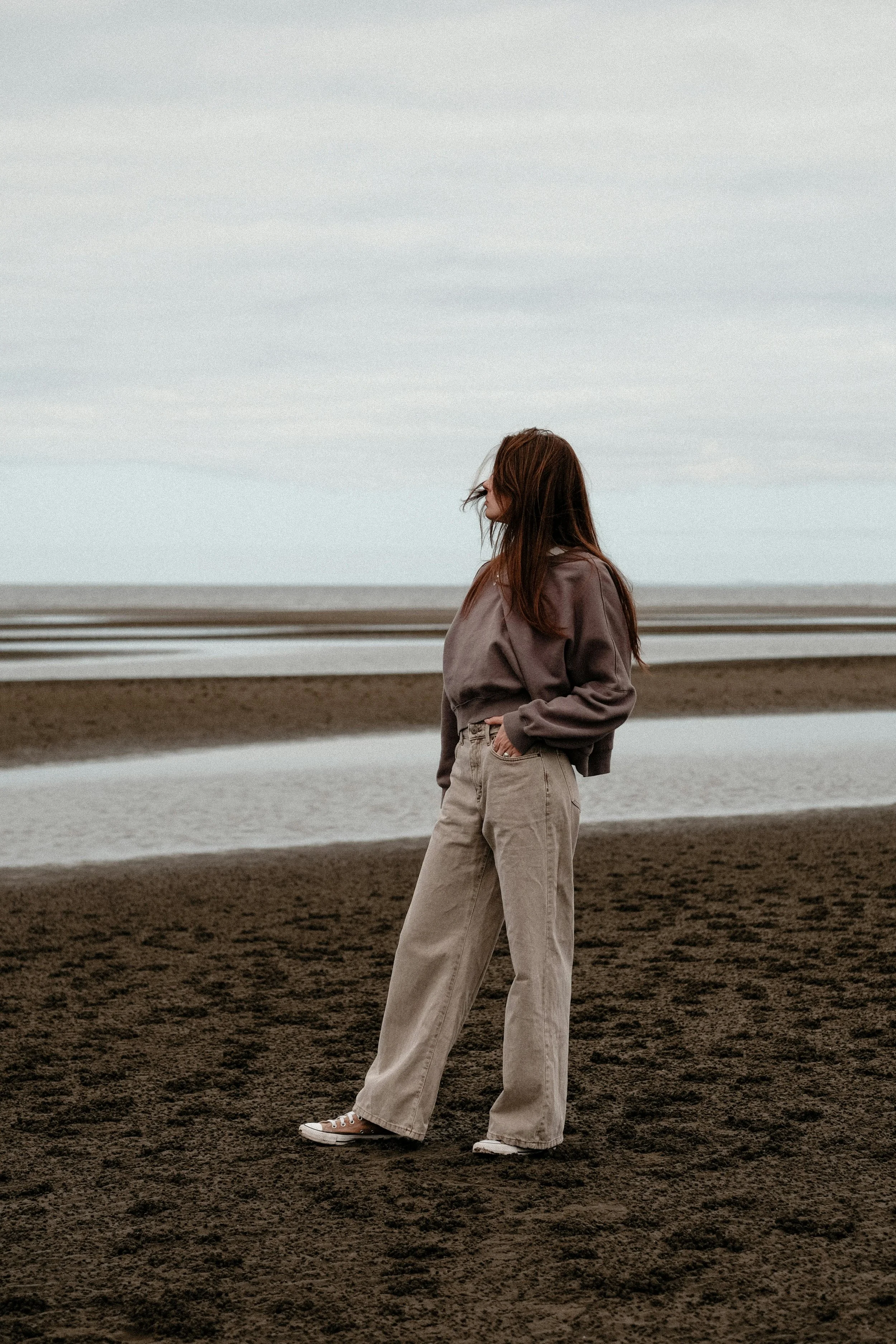 A woman with long brown hair standing on a dark sandy beach, wearing a brown sweatshirt, beige wide-leg pants, and sneakers, looking away towards the ocean under a cloudy sky.