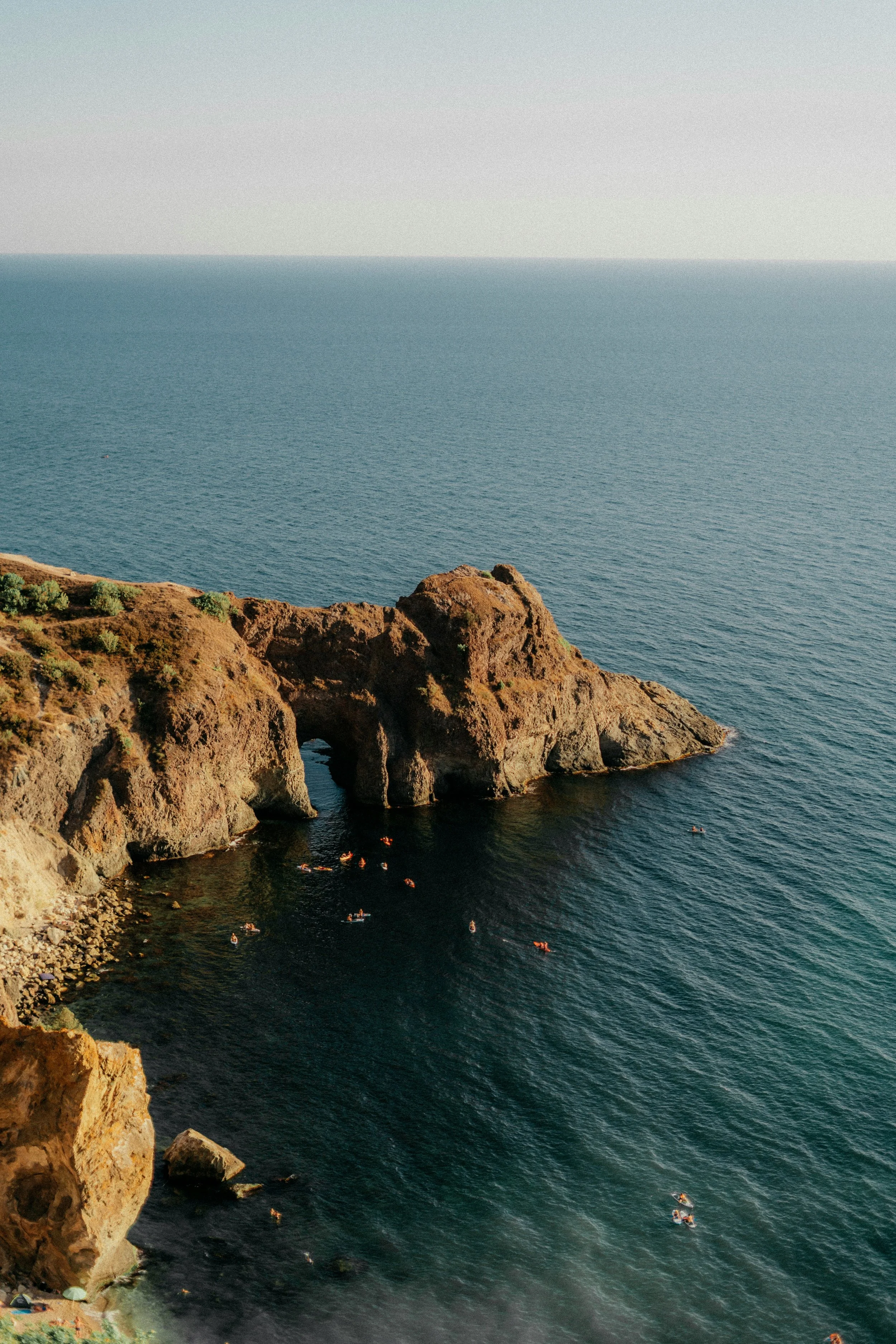 A coastal scene featuring a rocky cliff with a natural arch formation, overlooking the ocean with small boats in the water and a clear sky above.